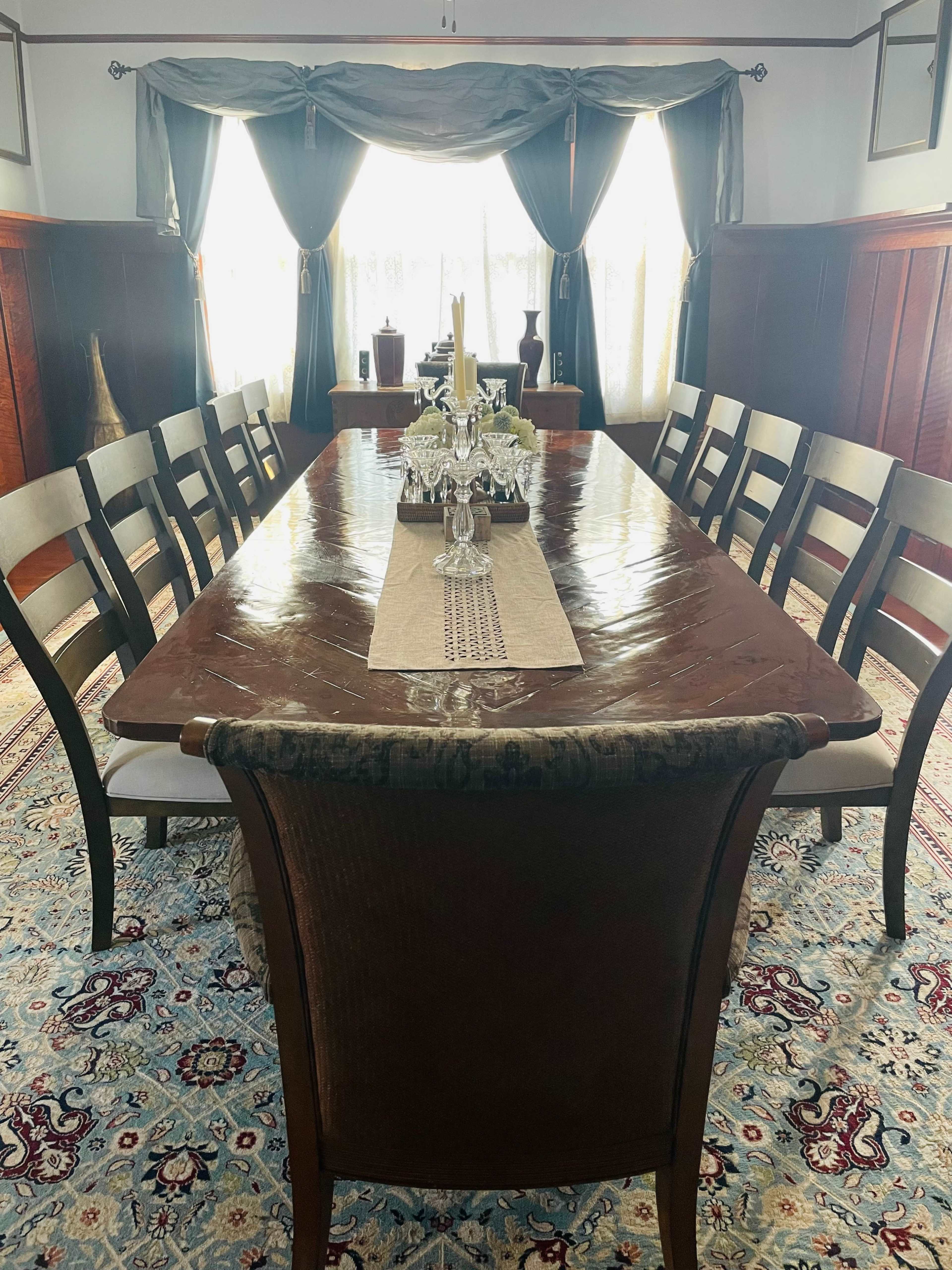 A long wooden dining table is set in a well-lit room with a chandelier and decorative curtains, surrounded by ten chairs.