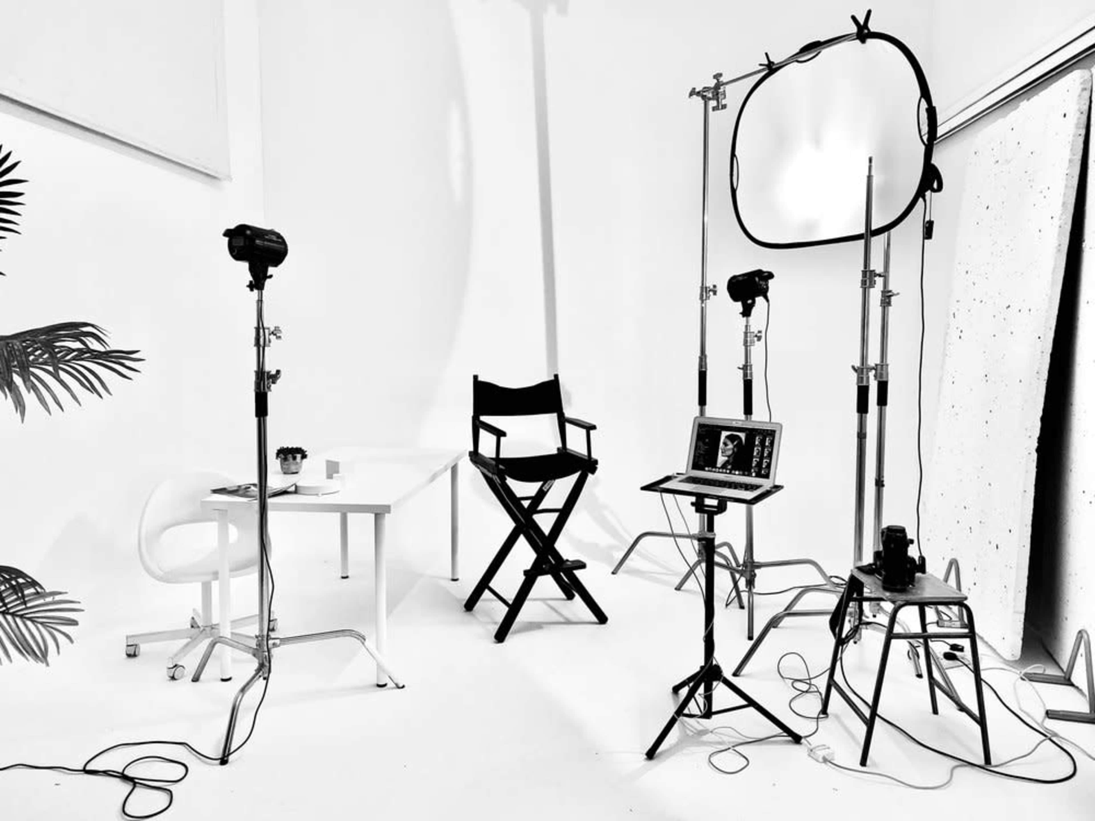A photography studio features a director's chair, a table with items, and several lighting equipment setups against a plain white background.