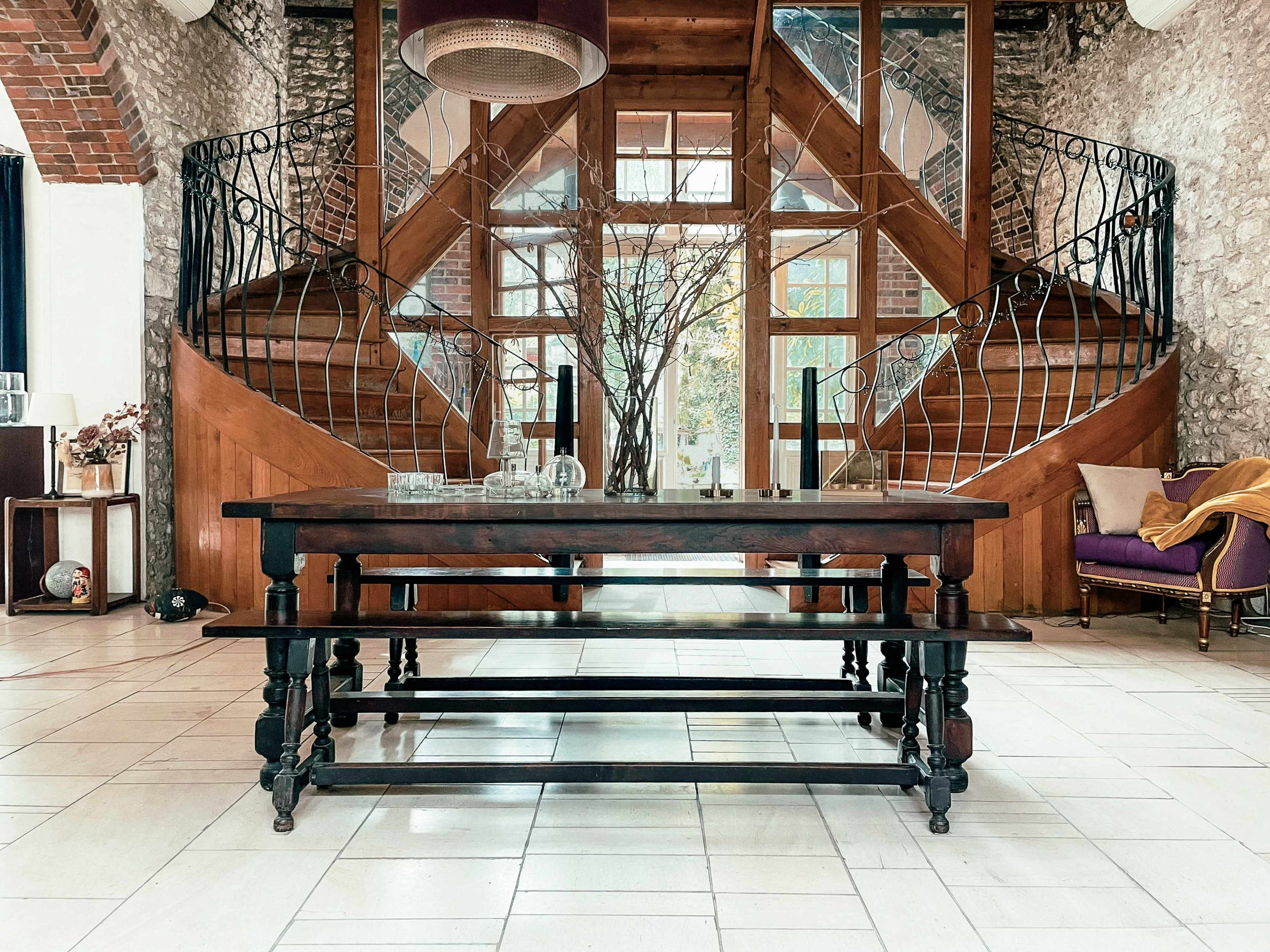 A wooden table is positioned in front of a large staircase with decorative railings, set in a room with stone walls and ample natural light.