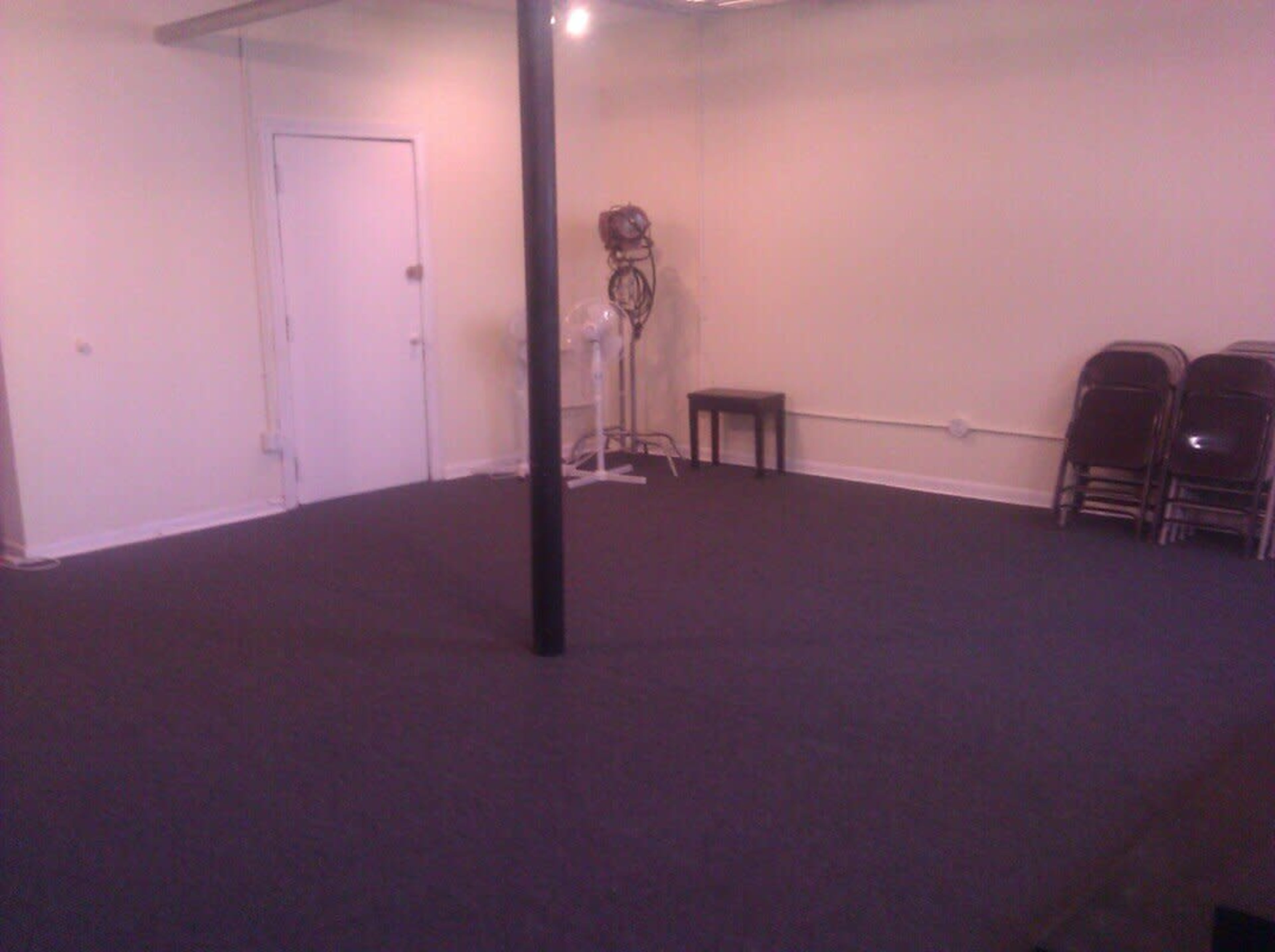 An empty room with a dark carpet, a single metal pole in the center, and a fan against the wall next to stacked metal chairs.