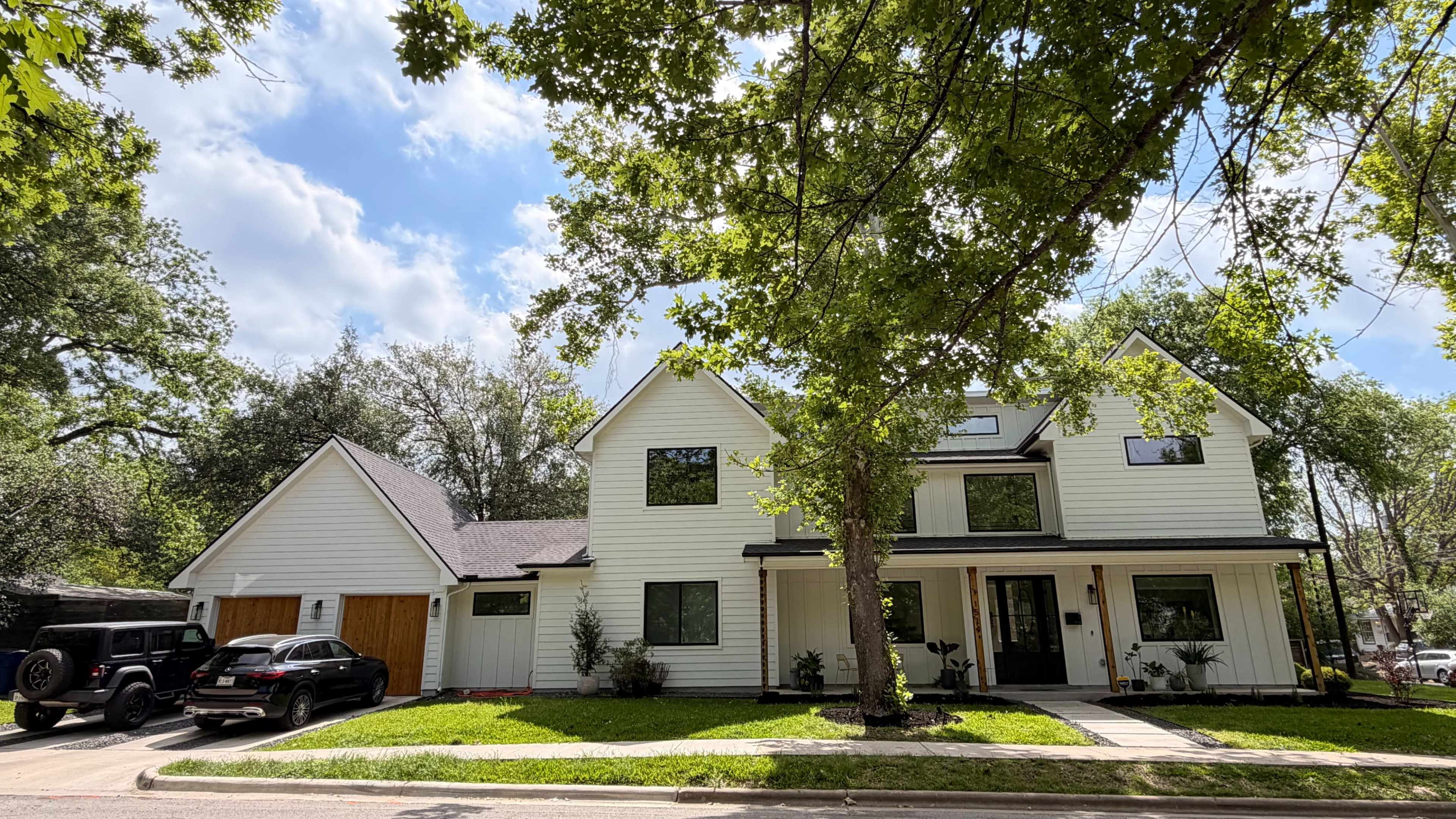 A two-story, modern farmhouse-style house with white exterior and a large front porch sits among trees on a residential street.