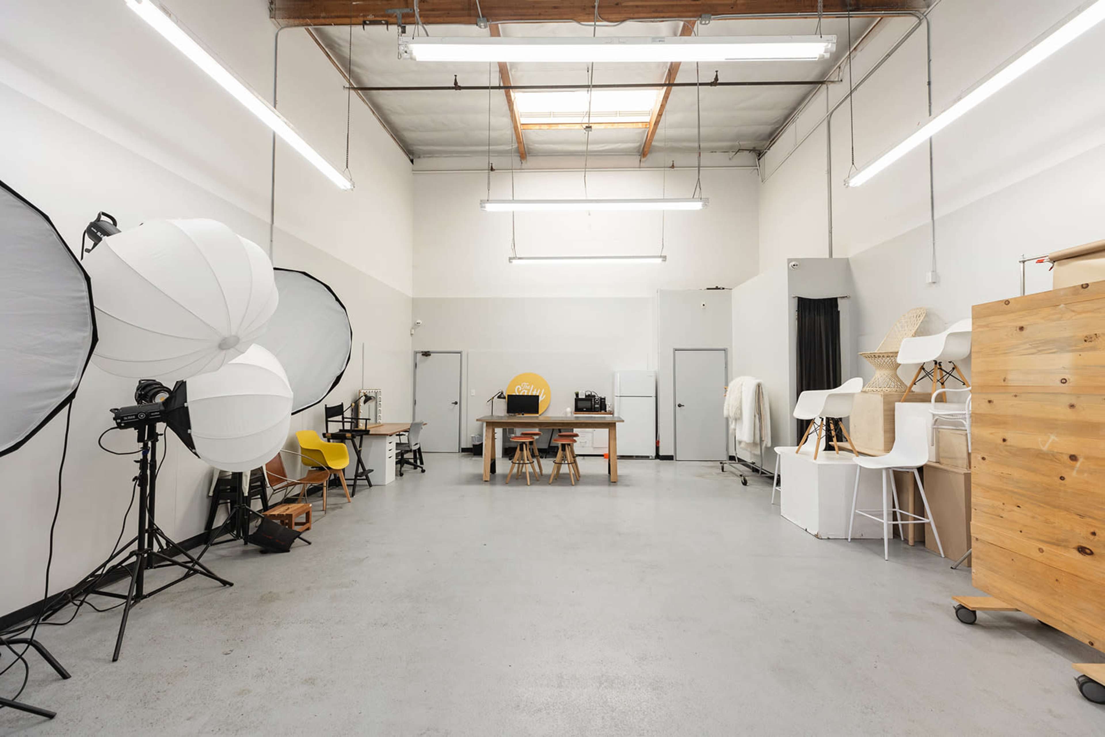 A spacious studio with photography equipment, including large softboxes, a table, chairs, and storage units against the walls.
