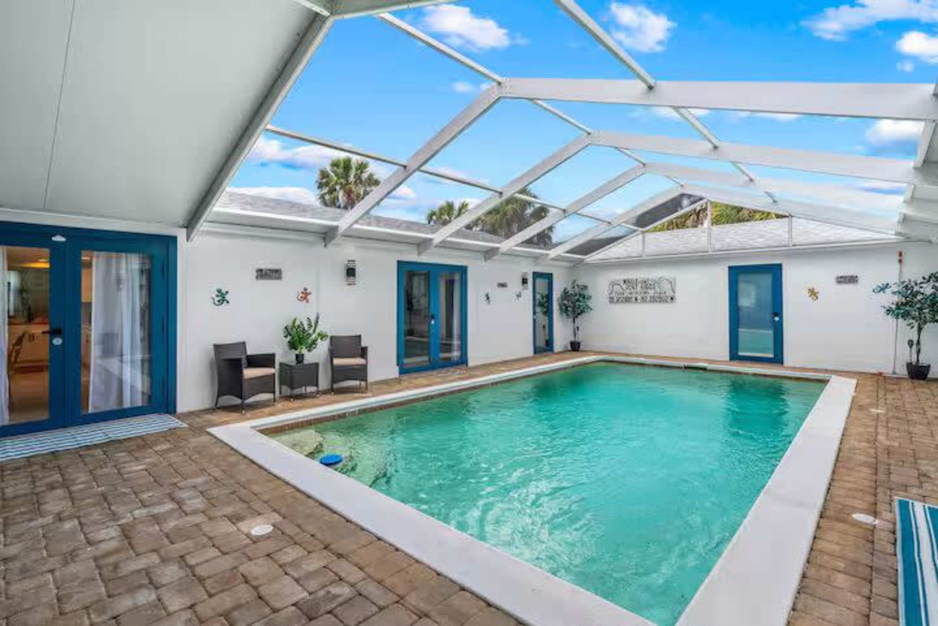 The image shows a rectangular swimming pool surrounded by brick flooring and a covered patio area with lounge chairs, situated inside a white-walled enclosure.