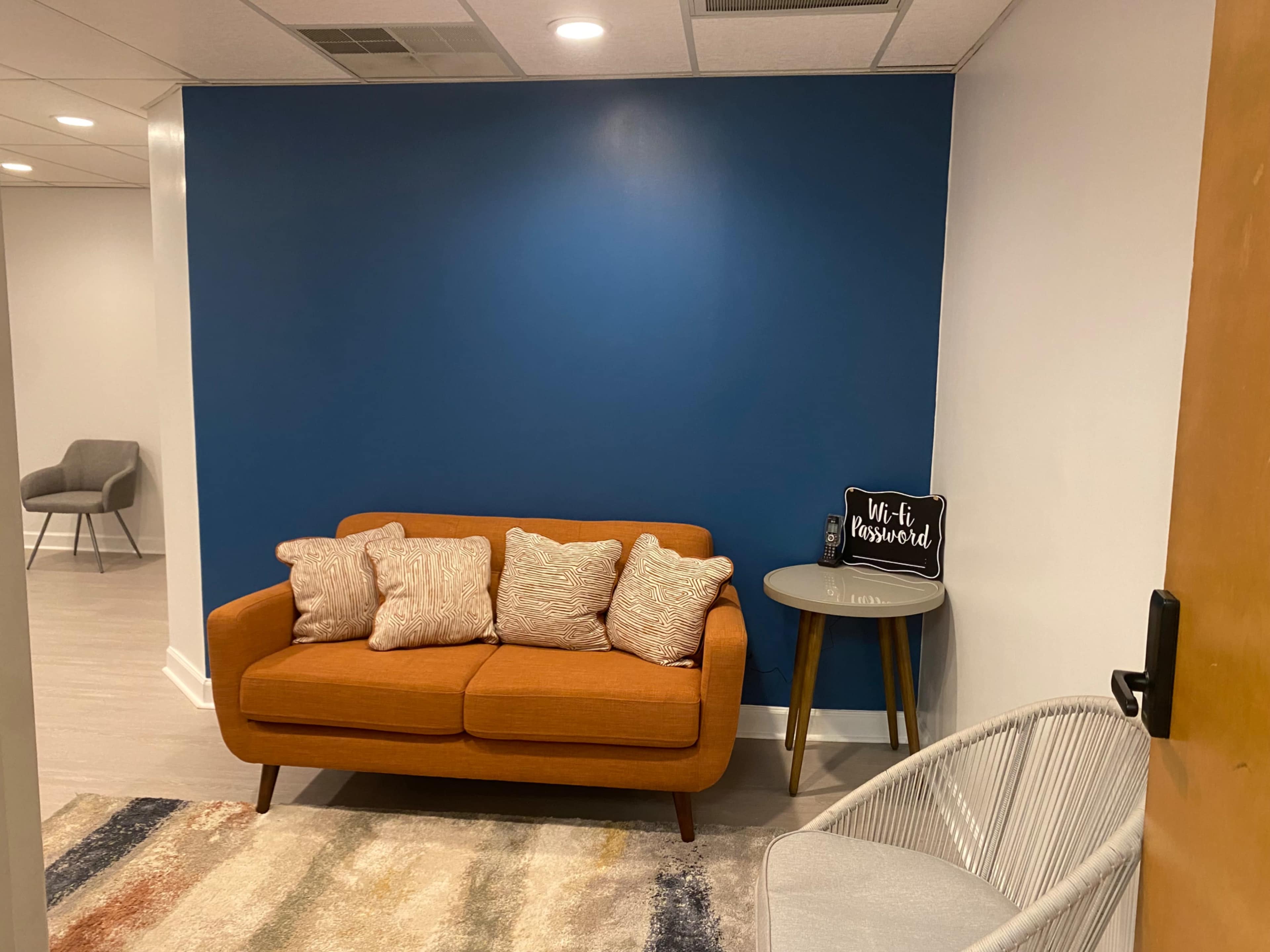 The image shows a small waiting area with an orange couch, a round side table, and a gray chair against a blue wall.