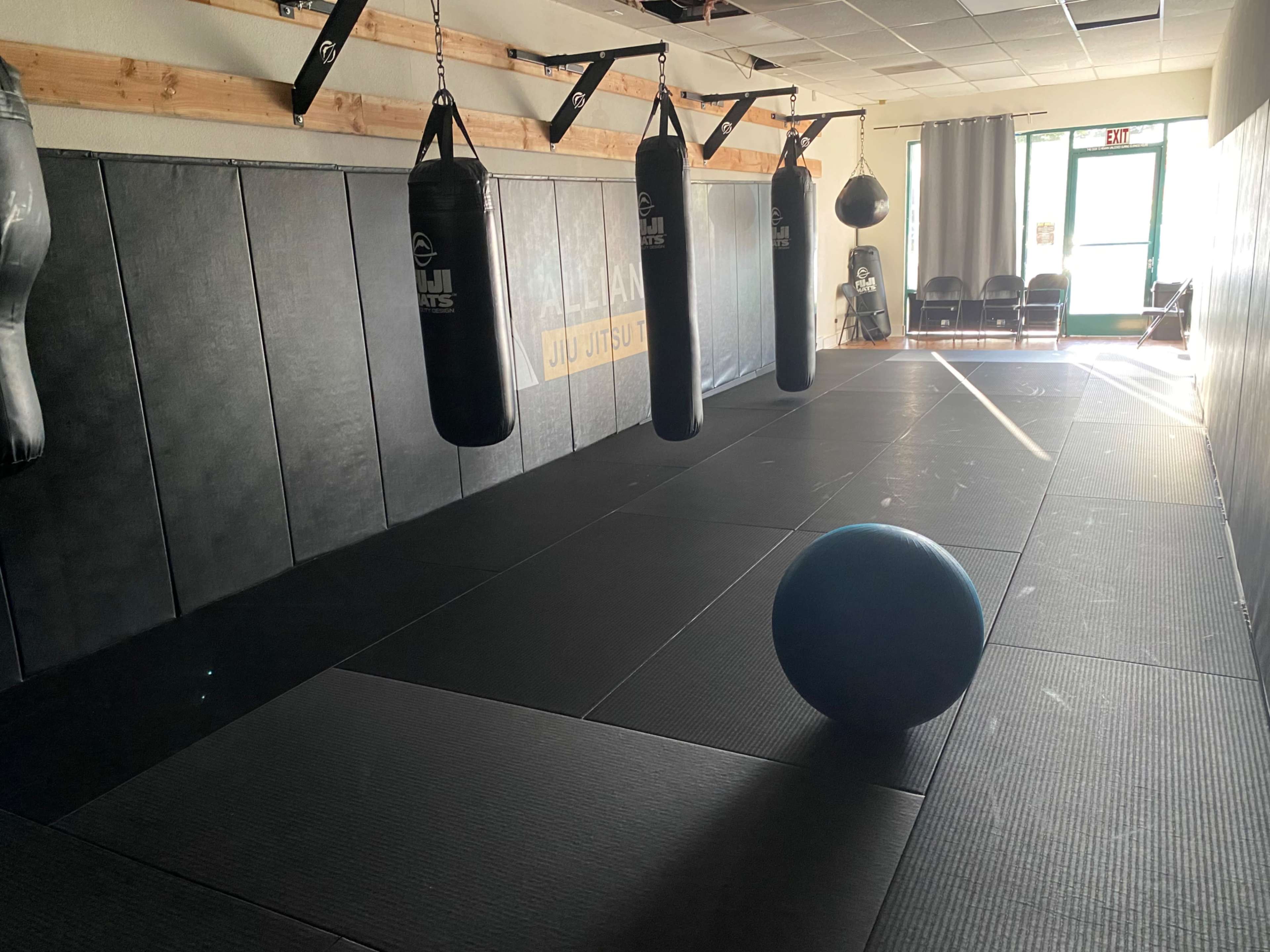 A sparsely furnished martial arts gym with black mats and hanging punching bags, featuring a large blue exercise ball positioned on the floor.