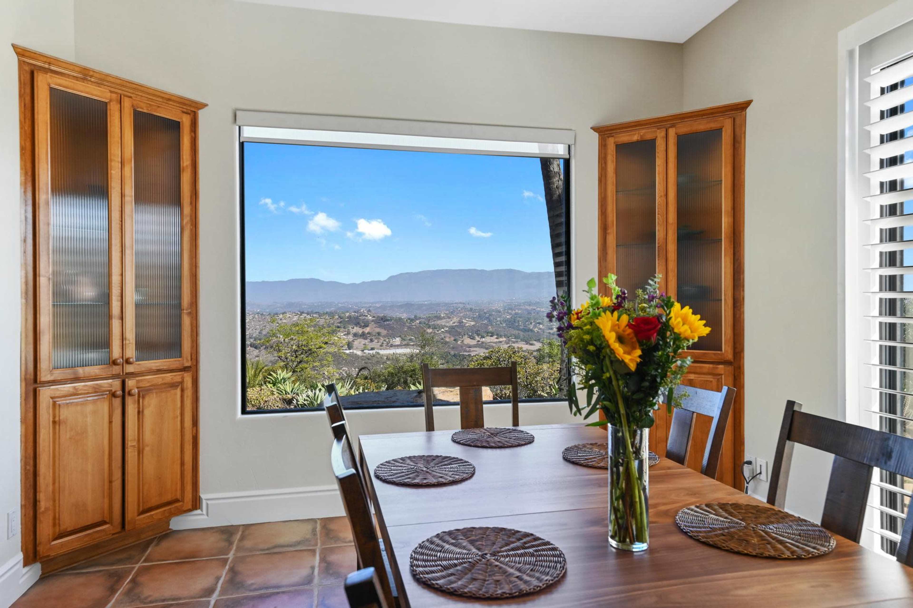 A dining area features a wooden table surrounded by chairs, with a large window showcasing a distant mountain view and sunlight streaming in.