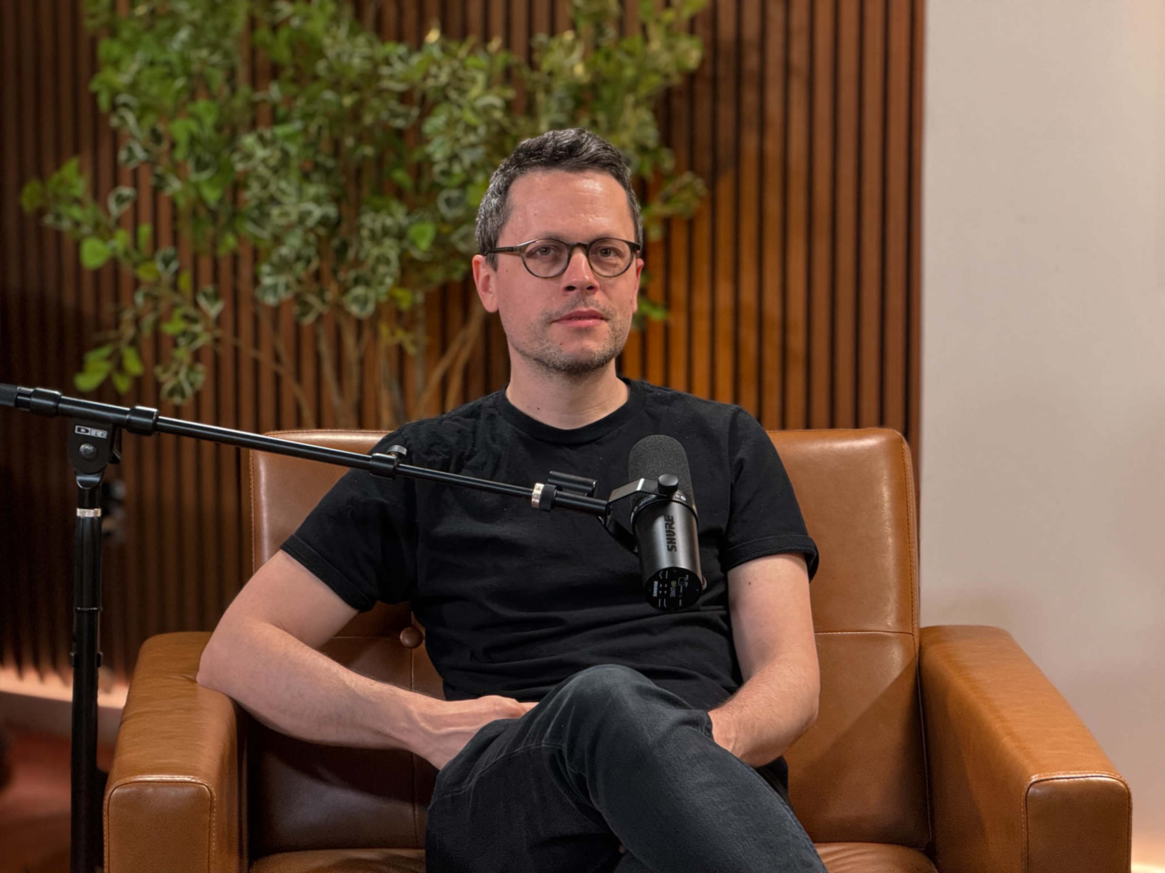A man with glasses sits in a brown leather chair, positioned near a microphone and a plant backdrop.