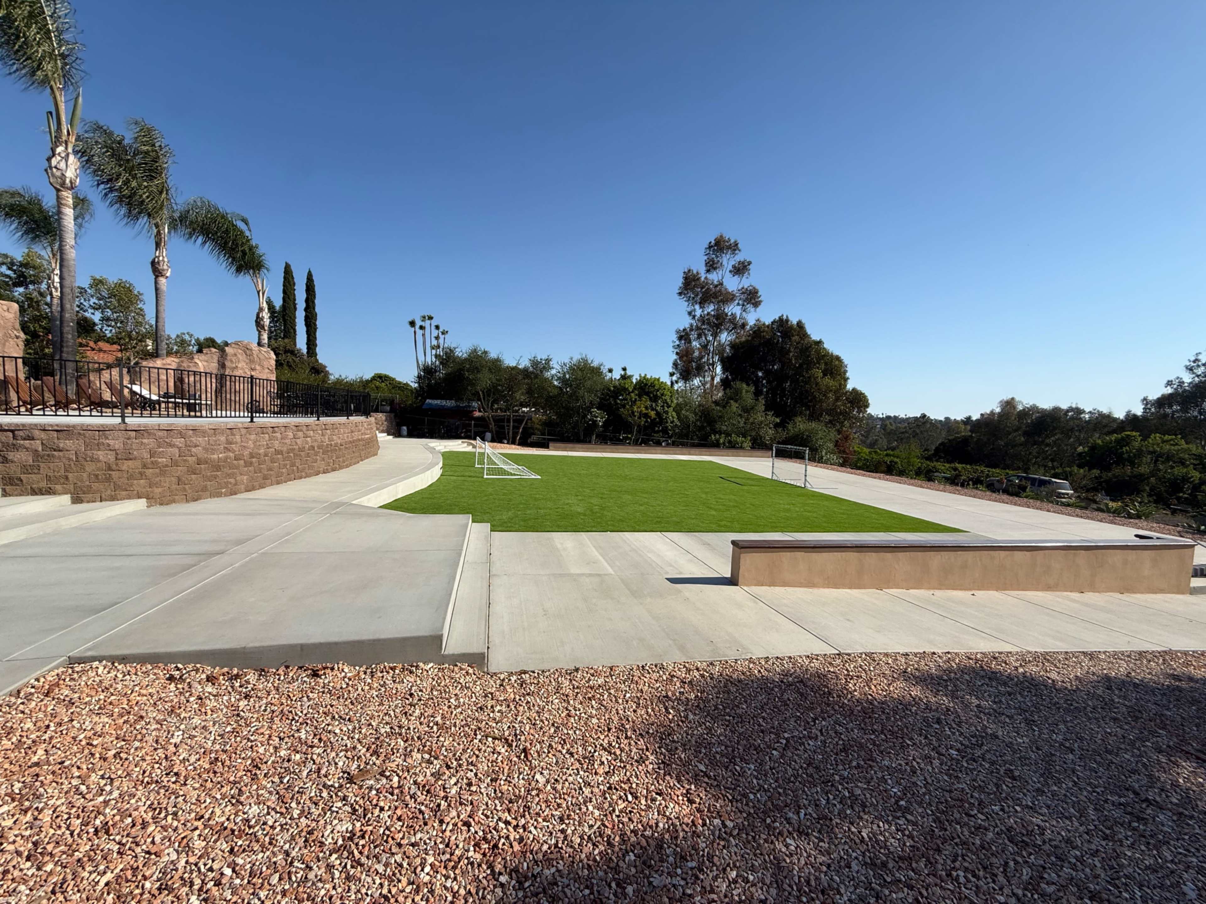 The image shows a spacious, flat outdoor area with a lawn, a concrete pathway, and two portable soccer goals, surrounded by landscaping and trees under a clear blue sky.