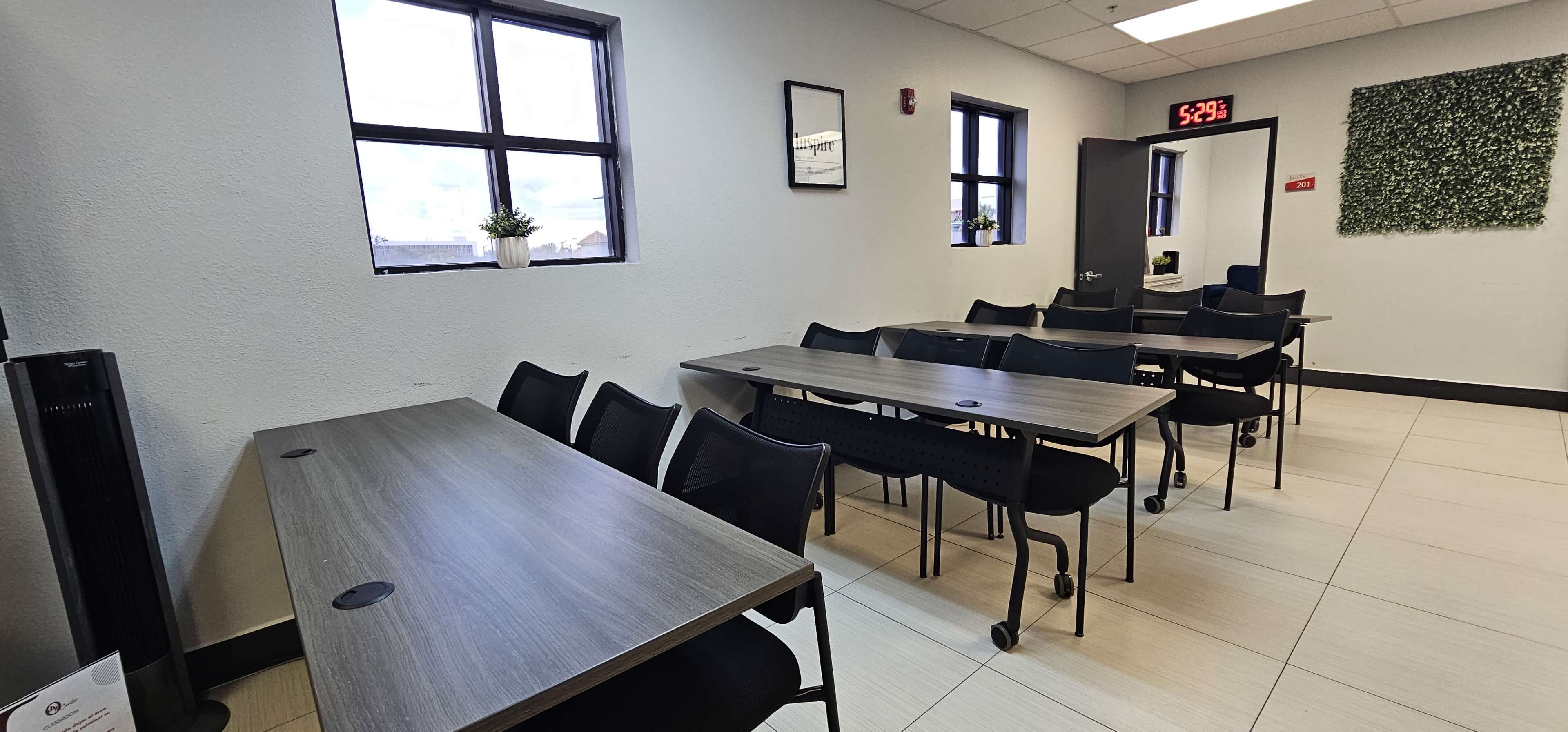 The image shows a spacious room with several tables and chairs arranged in rows, large windows allowing natural light, and green wall decor.