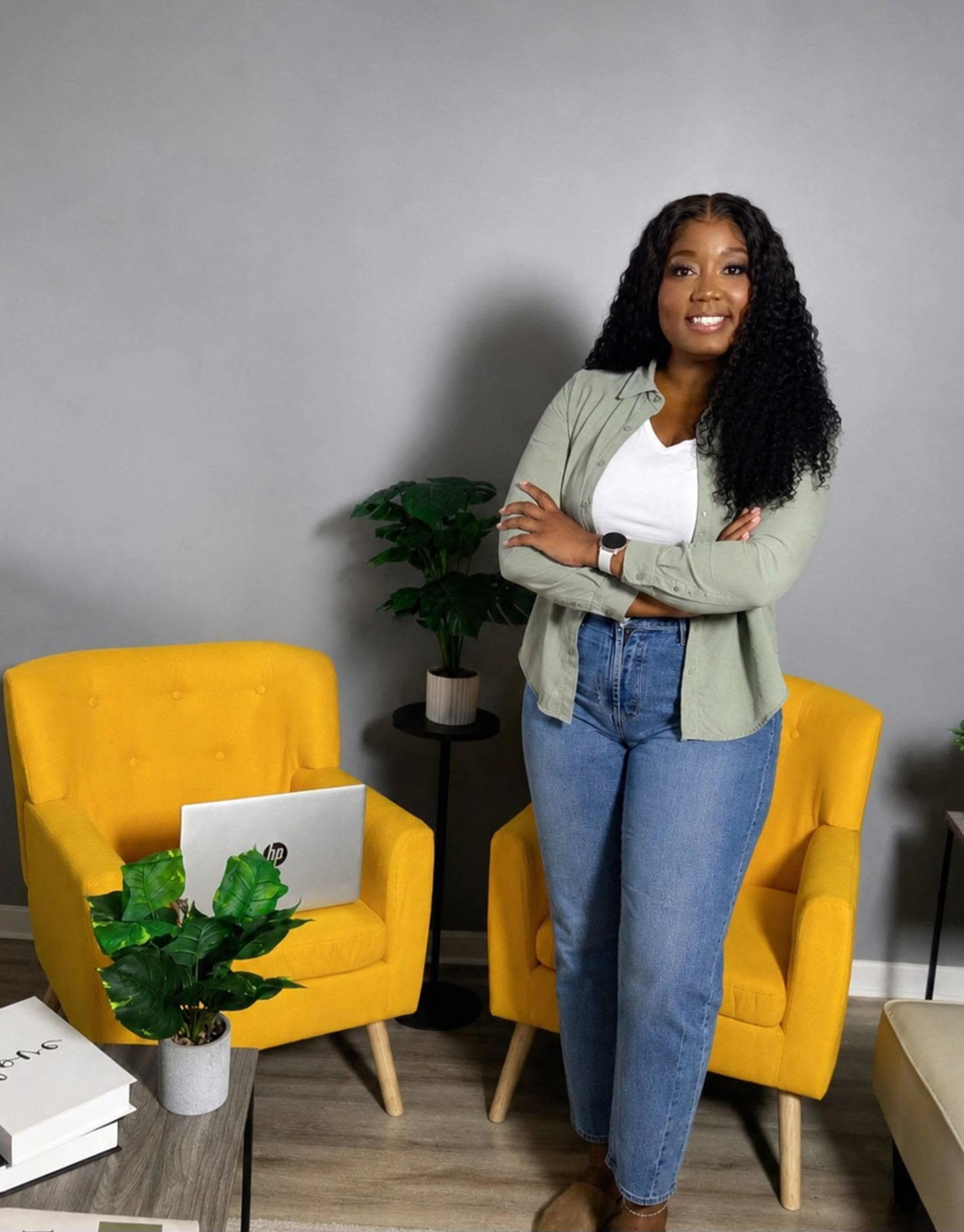 A person stands confidently with their arms crossed in a room featuring two yellow armchairs and a small table with a laptop and a plant.