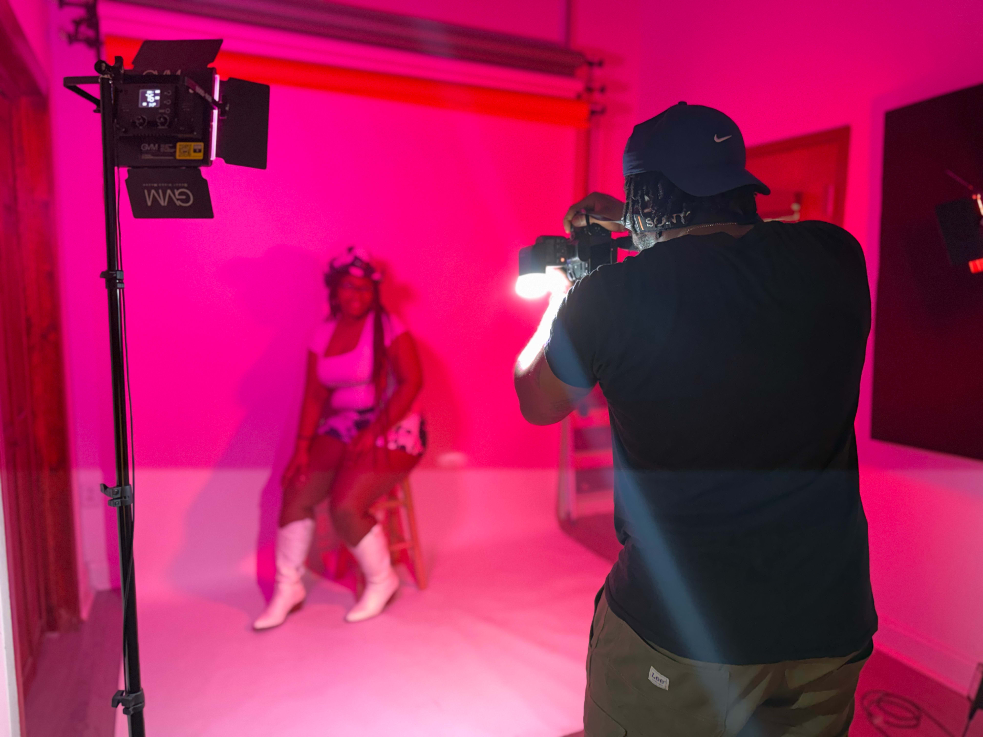 A photographer captures a model posed on a stool under pink lighting in a studio setting.
