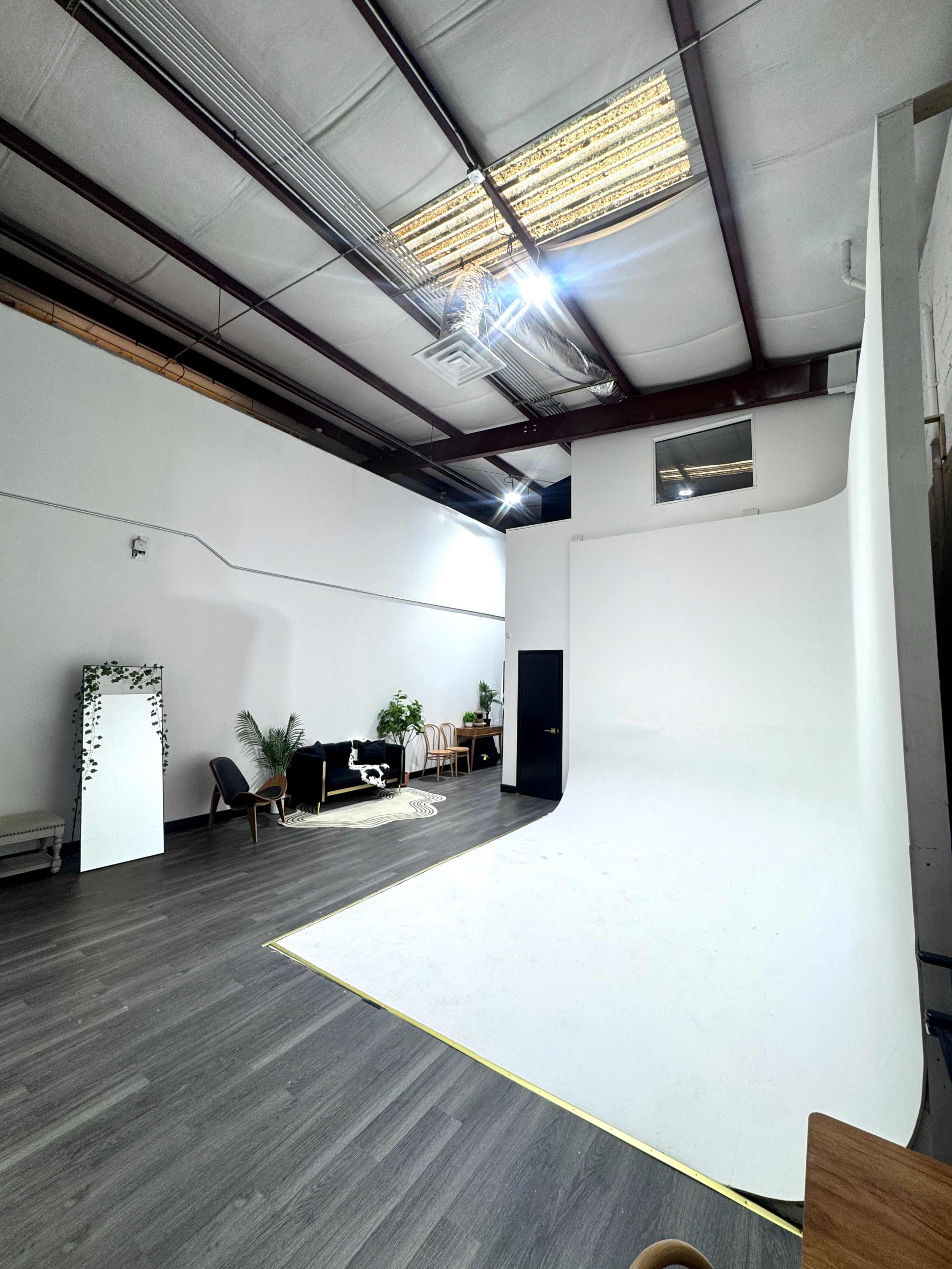 The image shows a spacious studio with a curved white backdrop, wooden flooring, and sparse furniture, including a sofa and chairs.