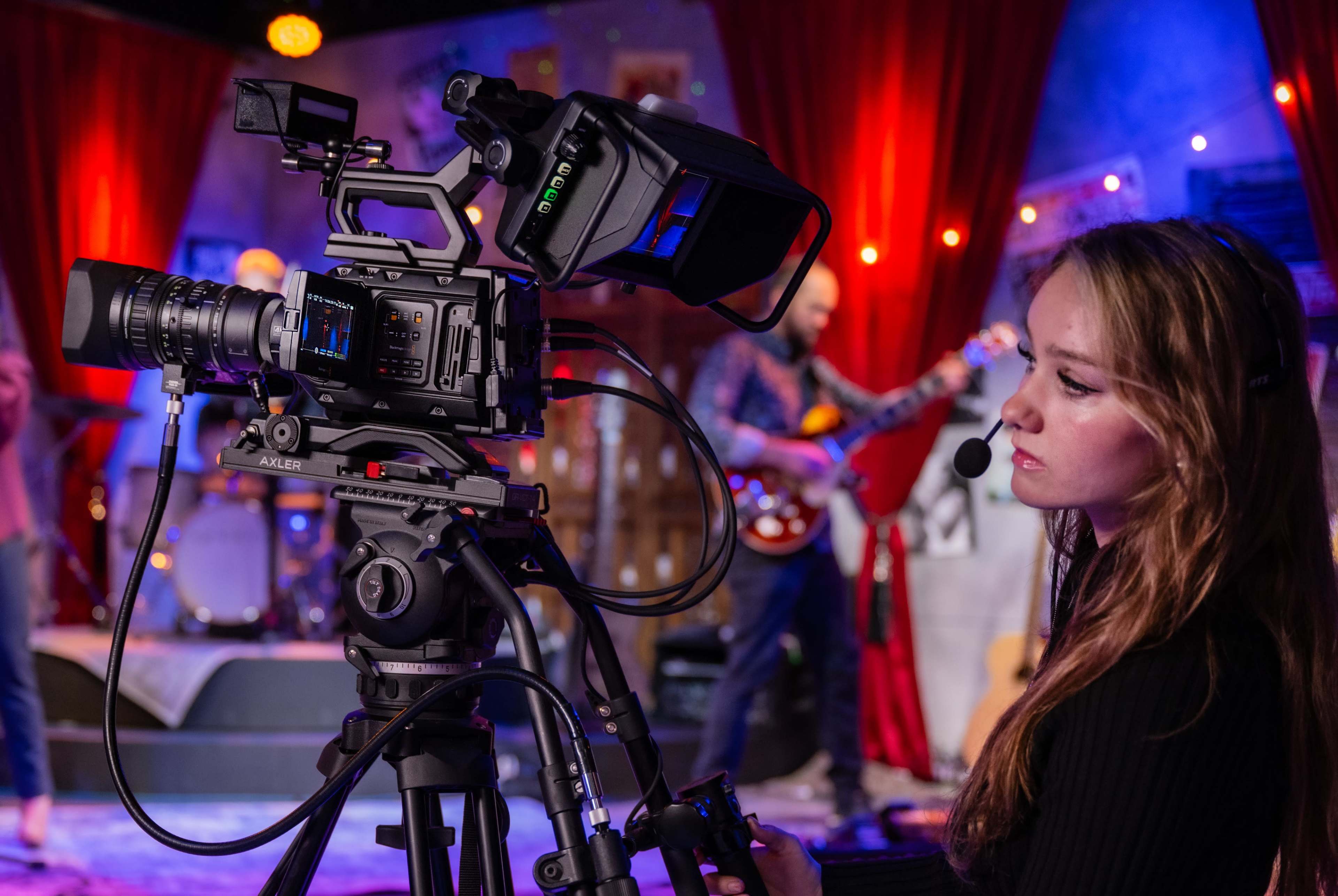 A camera operator monitors a professional video camera focused on a band performing on stage.