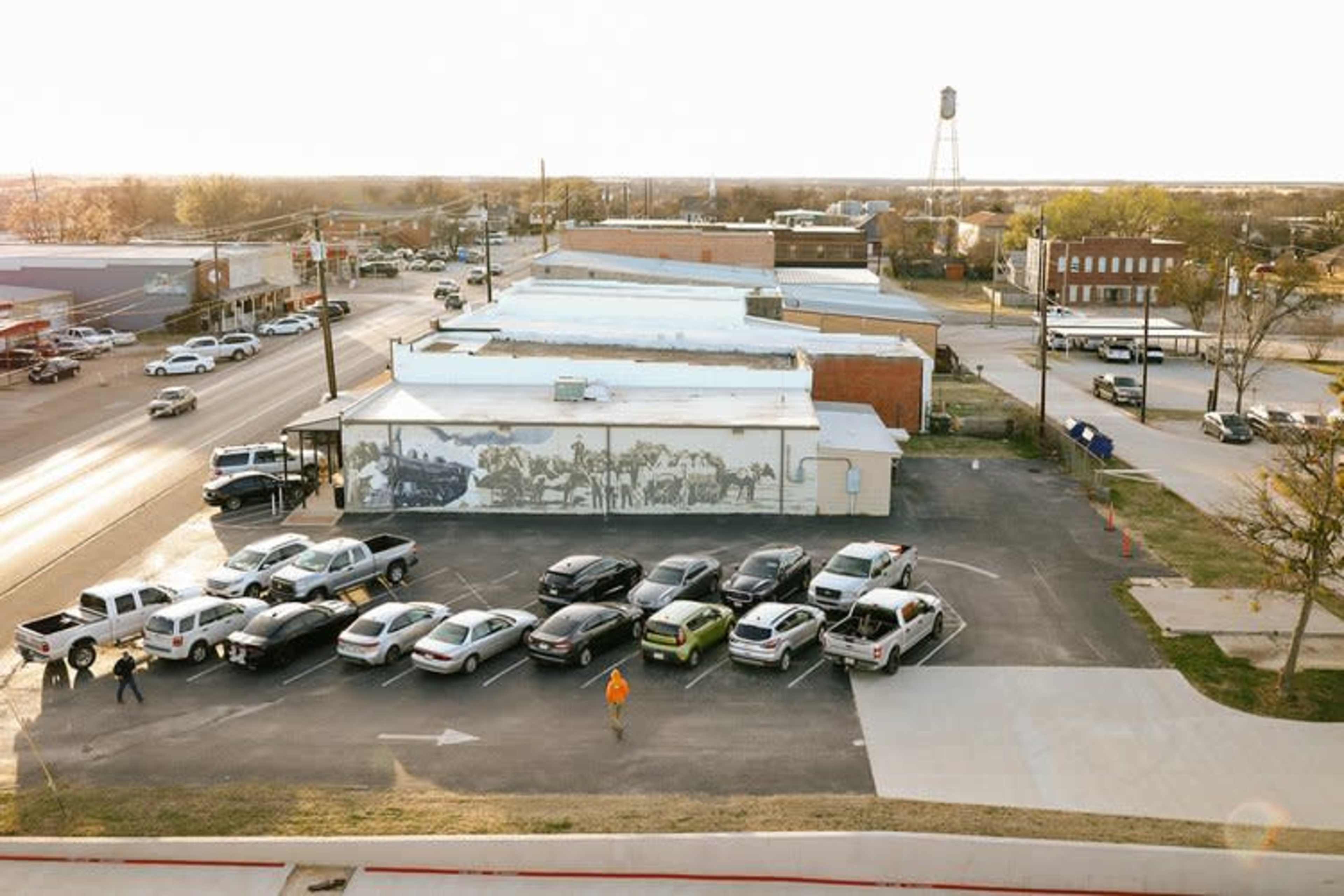 An aerial view of a parking lot filled with various parked cars next to a mural on a building and a street with several vehicles in the background.