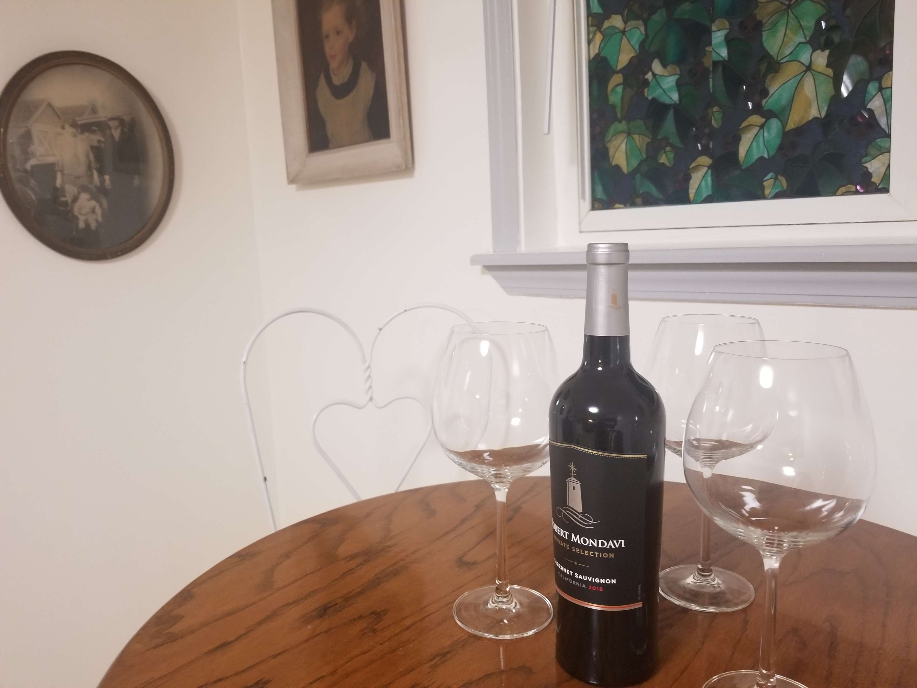 A bottle of wine and three wine glasses are placed on a wooden table in a room with two framed artworks and a window showing green leaves.
