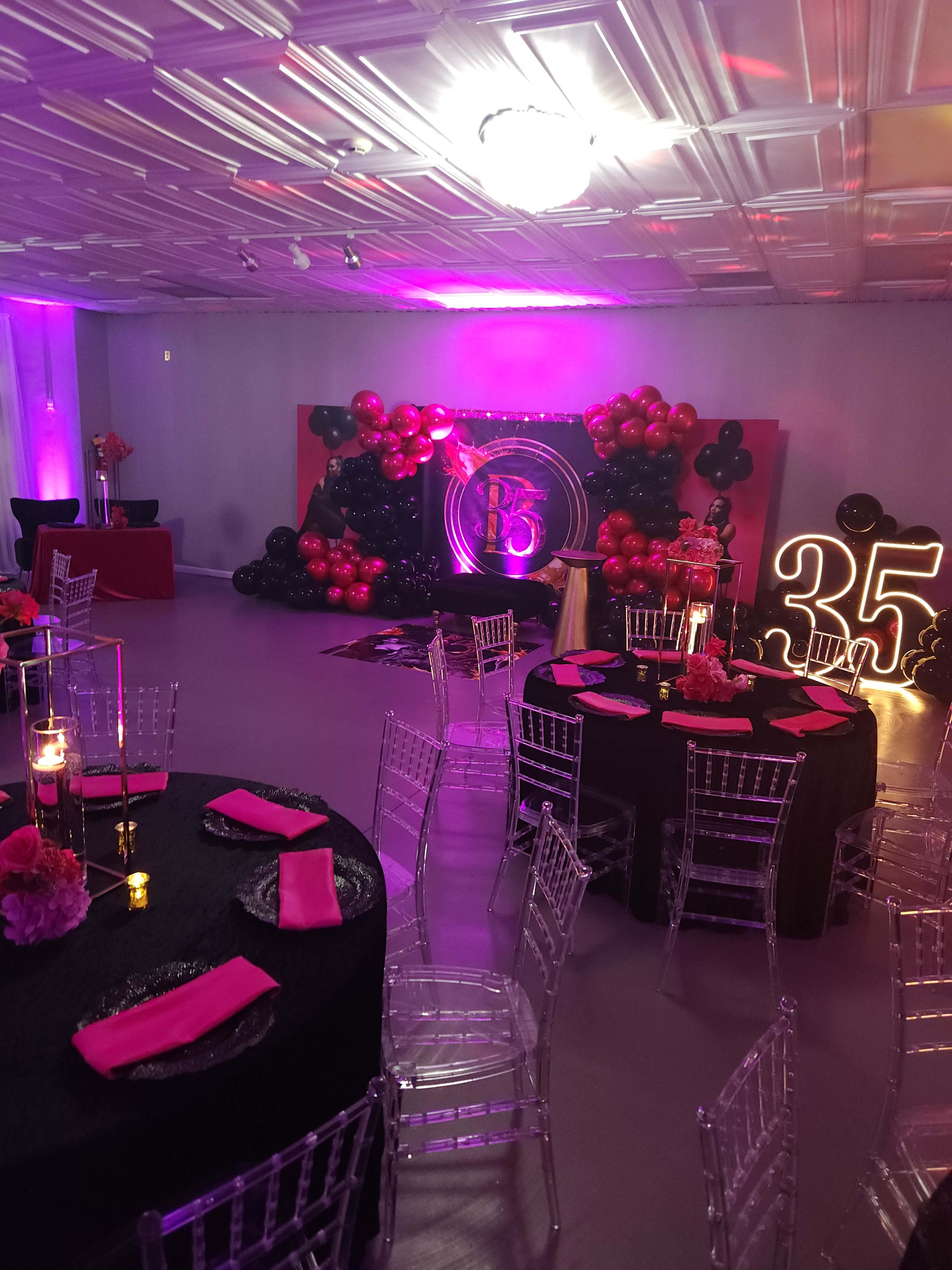 The image shows a decorated event space with black tables, pink napkins, and a backdrop featuring balloons and a glowing "35" for a celebration.