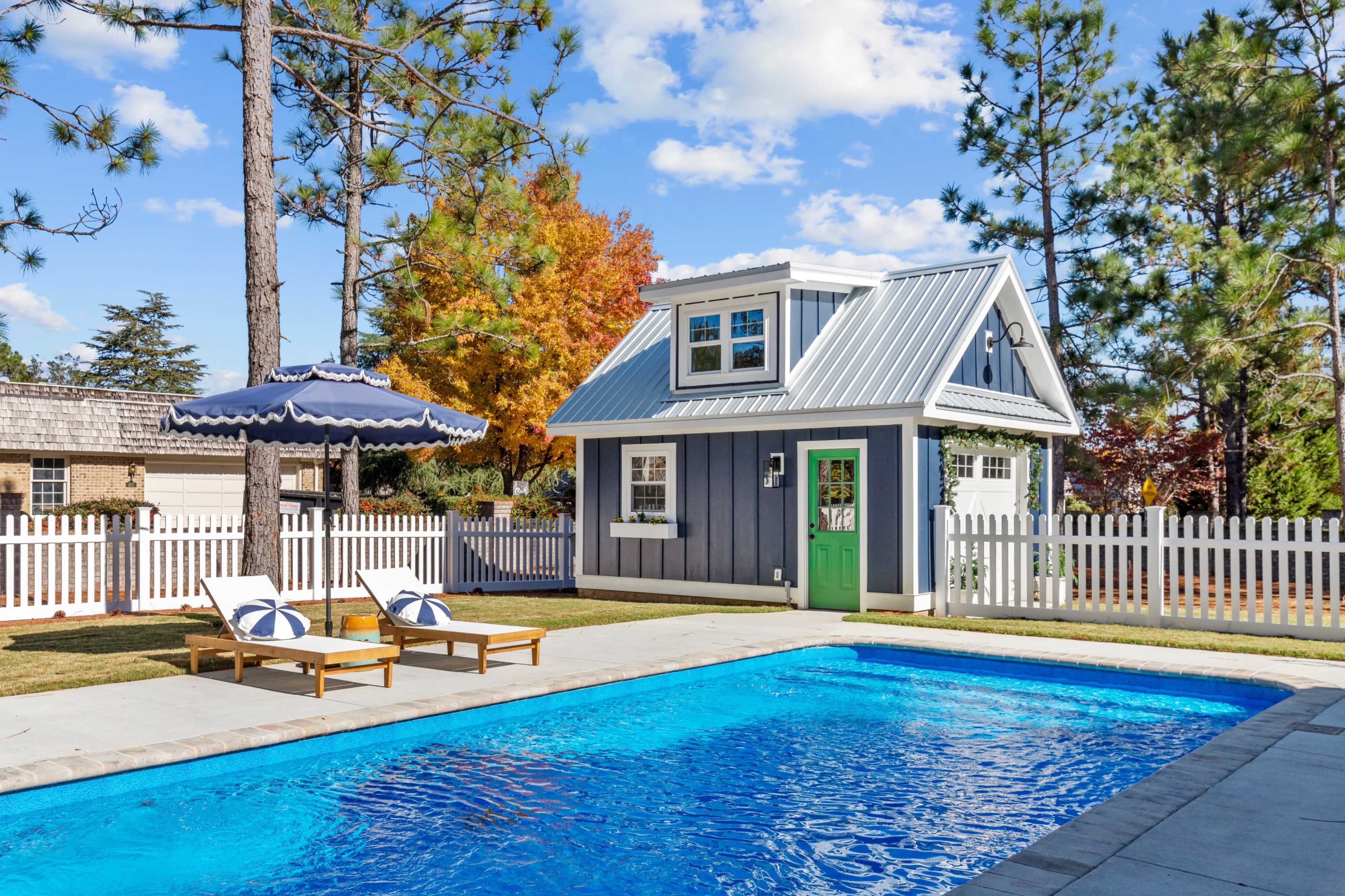 A backyard scene features a blue swimming pool in front of a small gray and white cottage, surrounded by tall trees and a white picket fence.