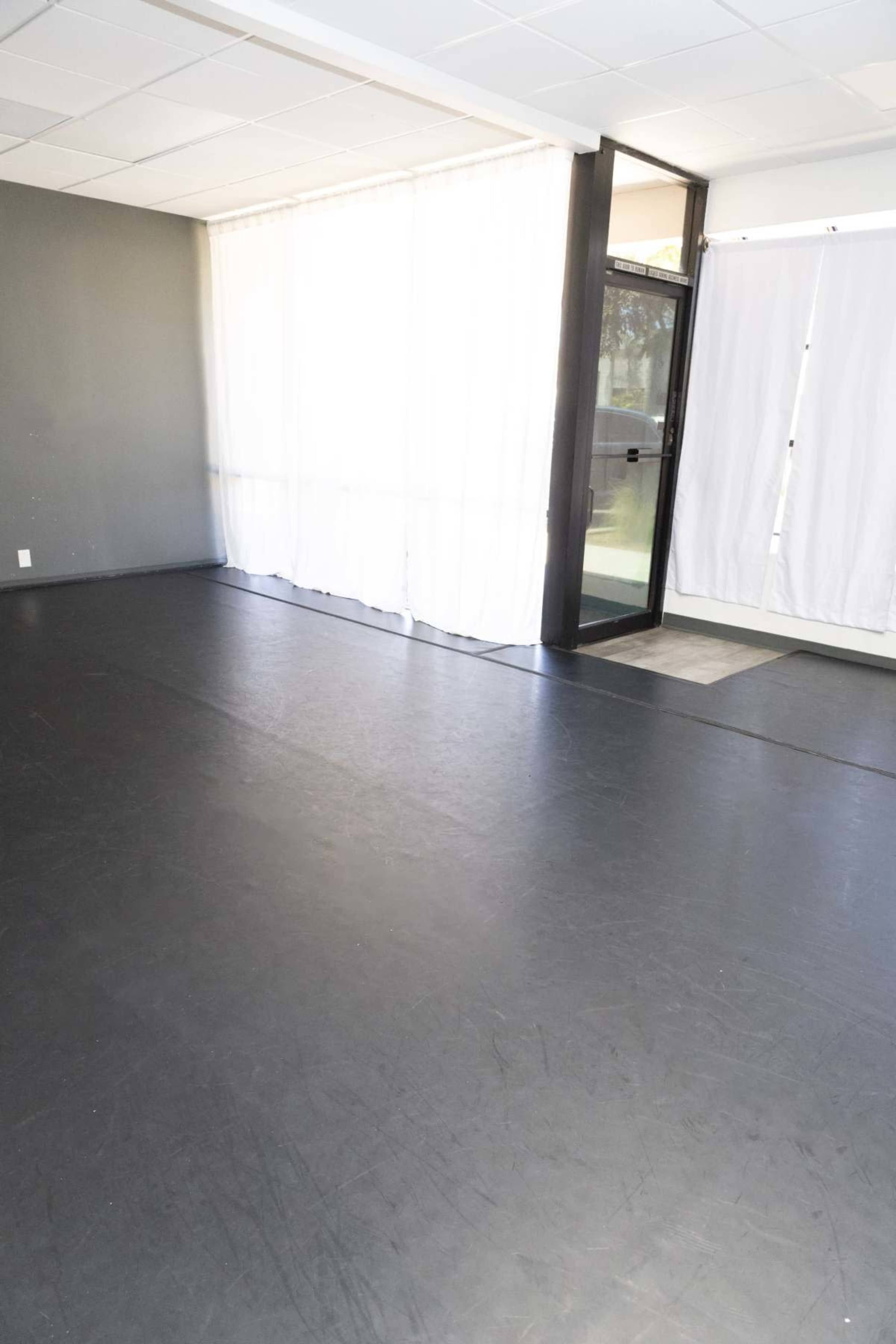 The image shows a spacious, empty room with a black floor, large windows covered by sheer white curtains, and a glass door leading outside.