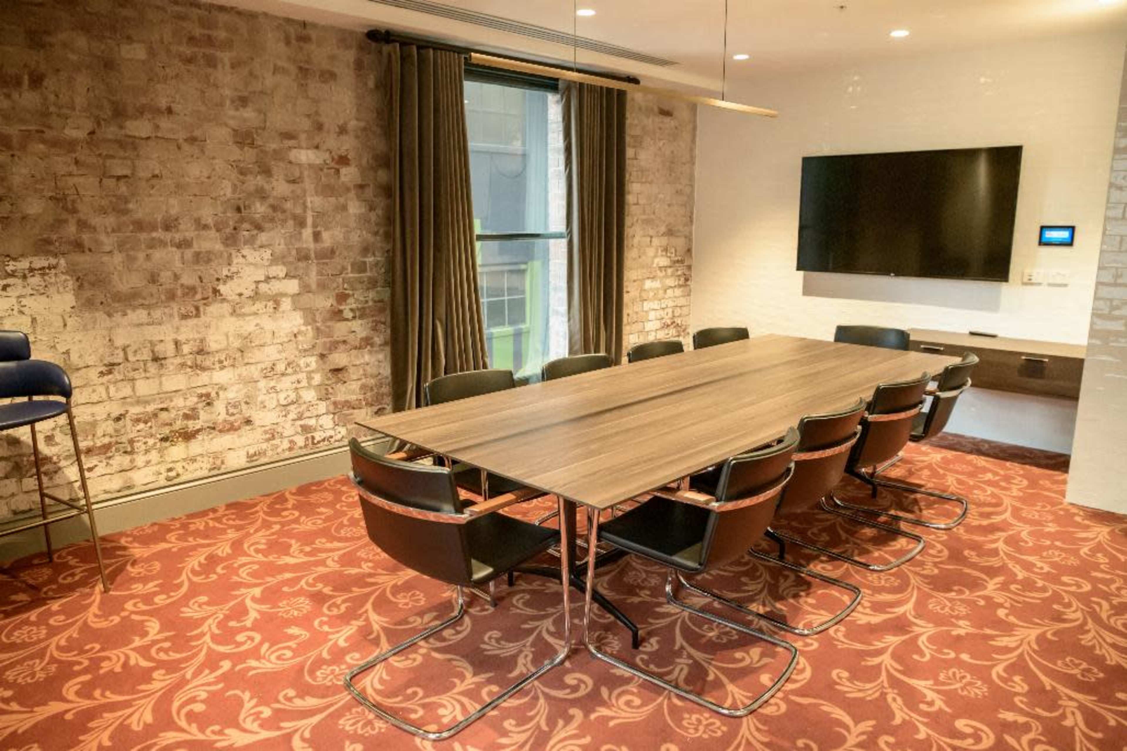 A conference room features a long wooden table surrounded by black chairs, with exposed brick walls and a wall-mounted television.