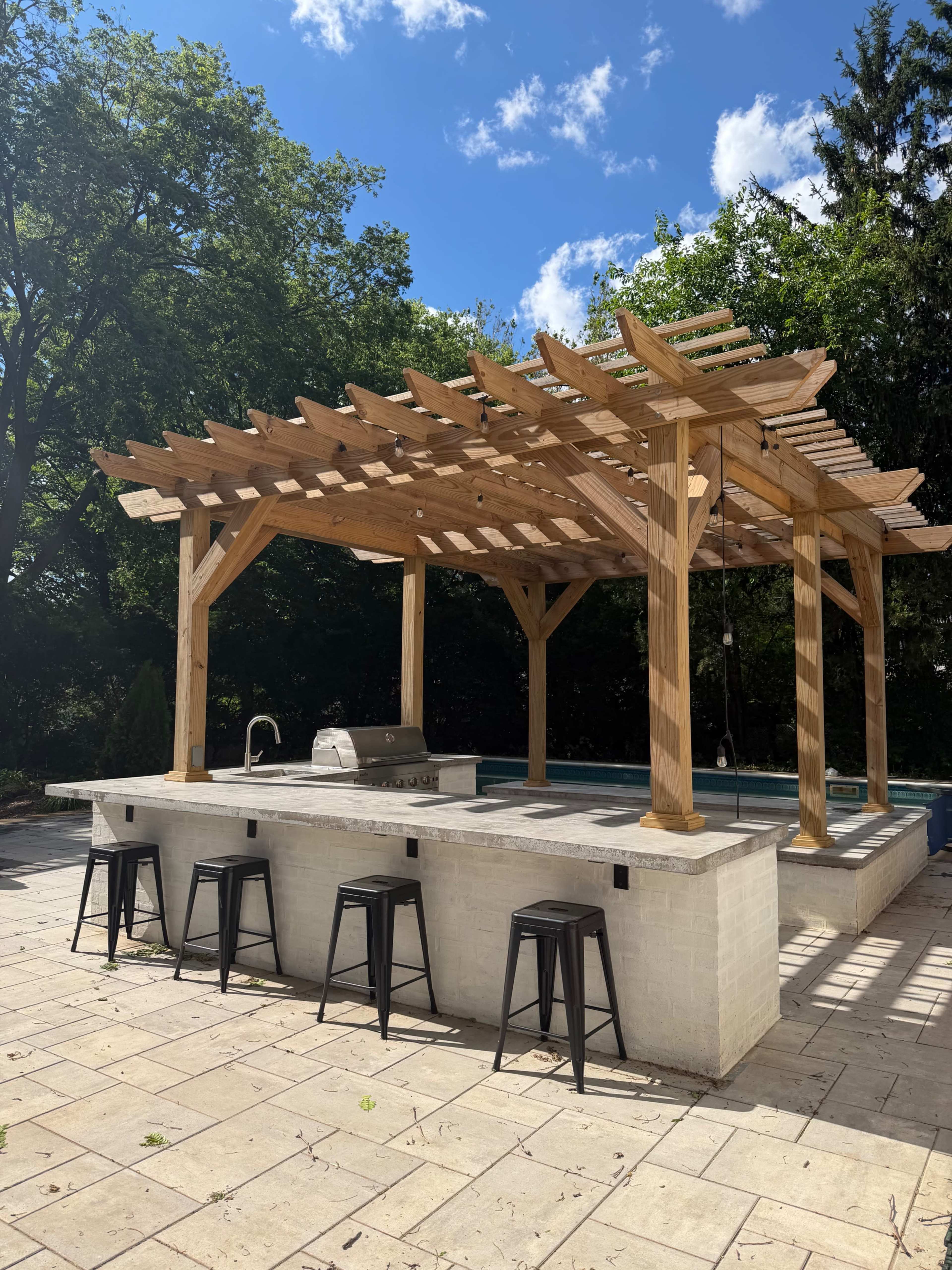 The image shows a wooden pergola over an outdoor kitchen area with a grill, surrounded by patio seating and a swimming pool in the background.