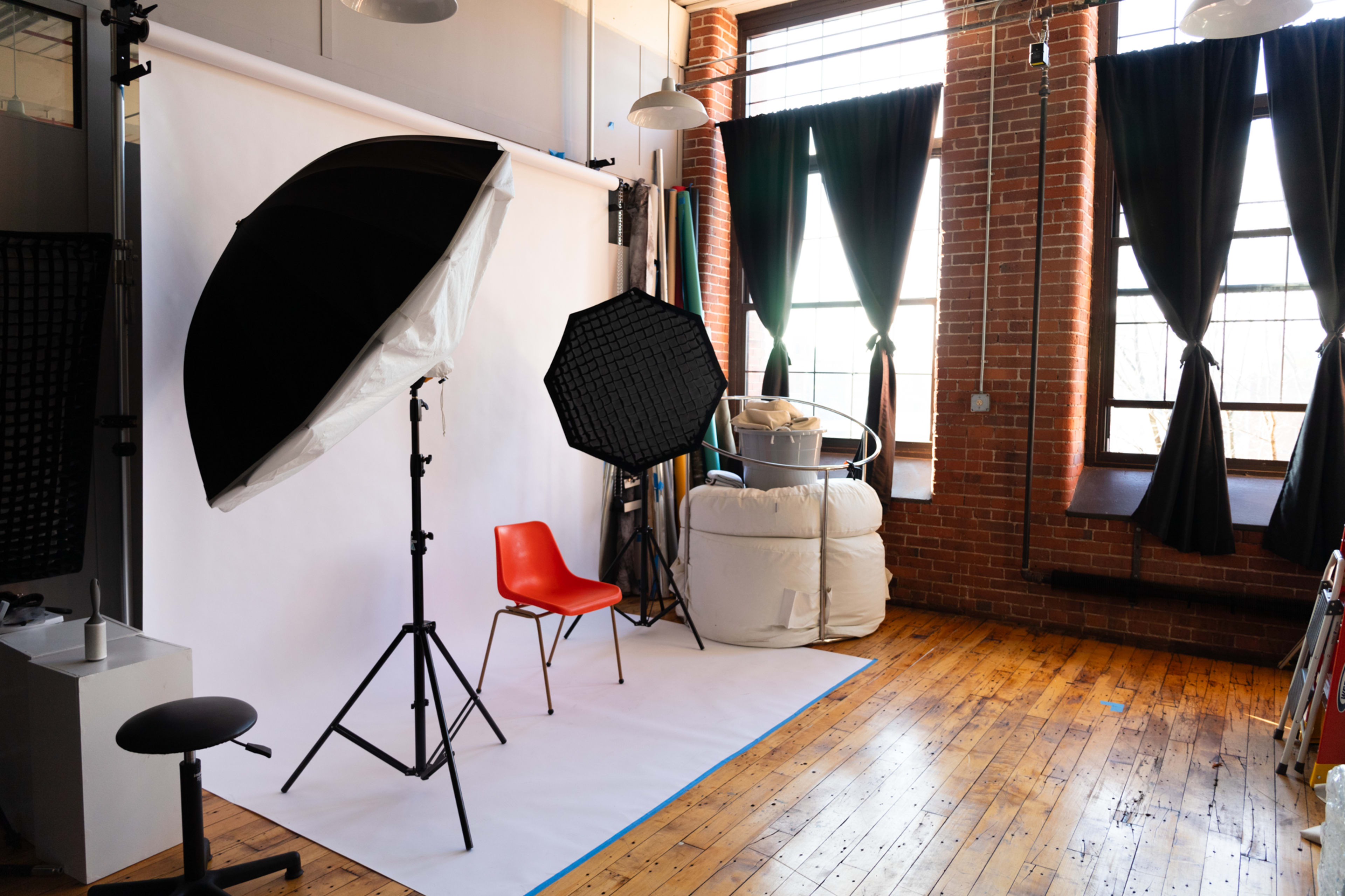 A photography studio with a white backdrop, a red chair, and various lighting equipment, set against a backdrop of large windows and brick walls.