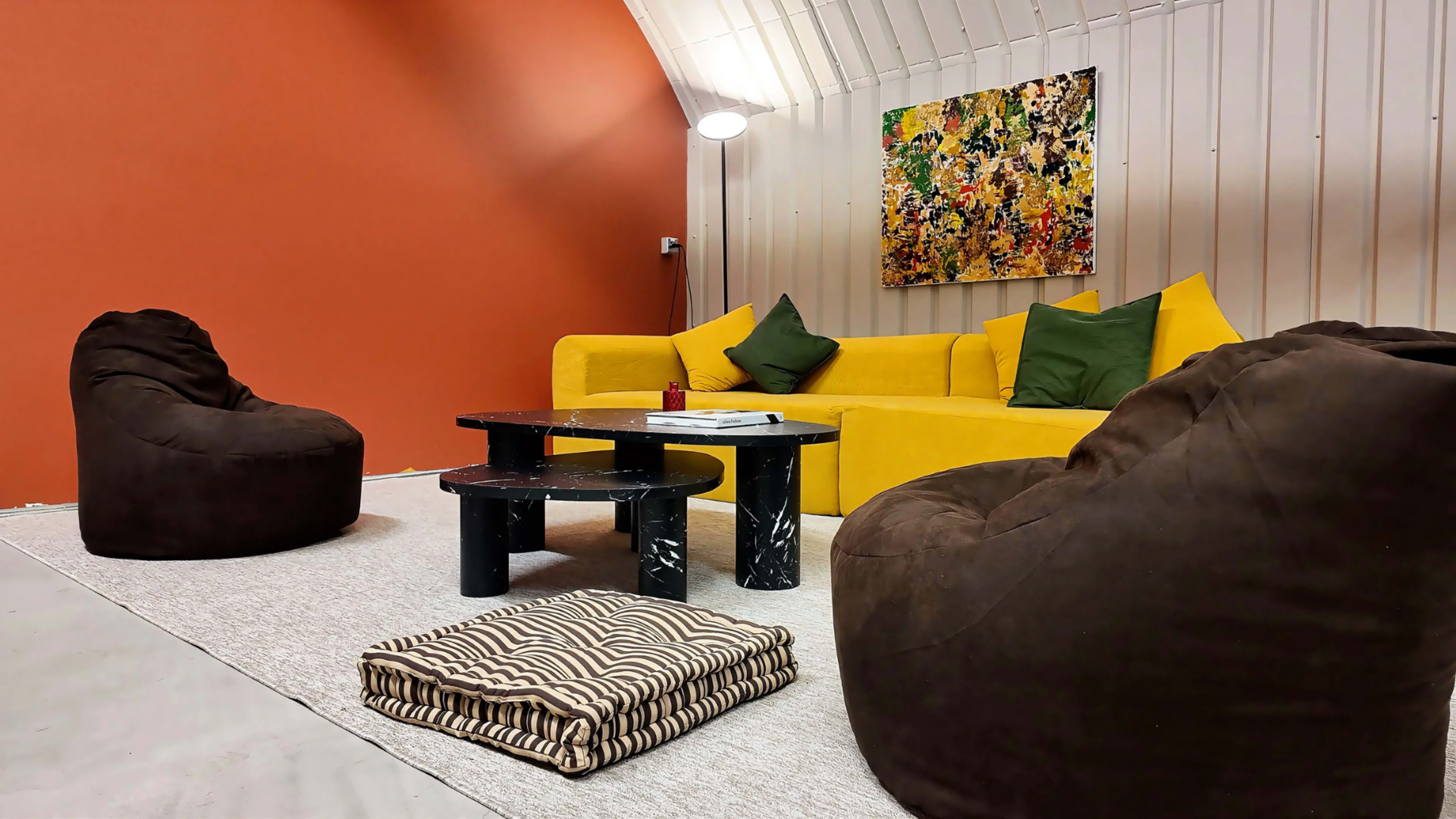 The scene shows a cozy living area with a yellow couch, two brown bean bag chairs, a small black coffee table, and a striped floor cushion, set against an orange wall and decorated with an abstract painting.