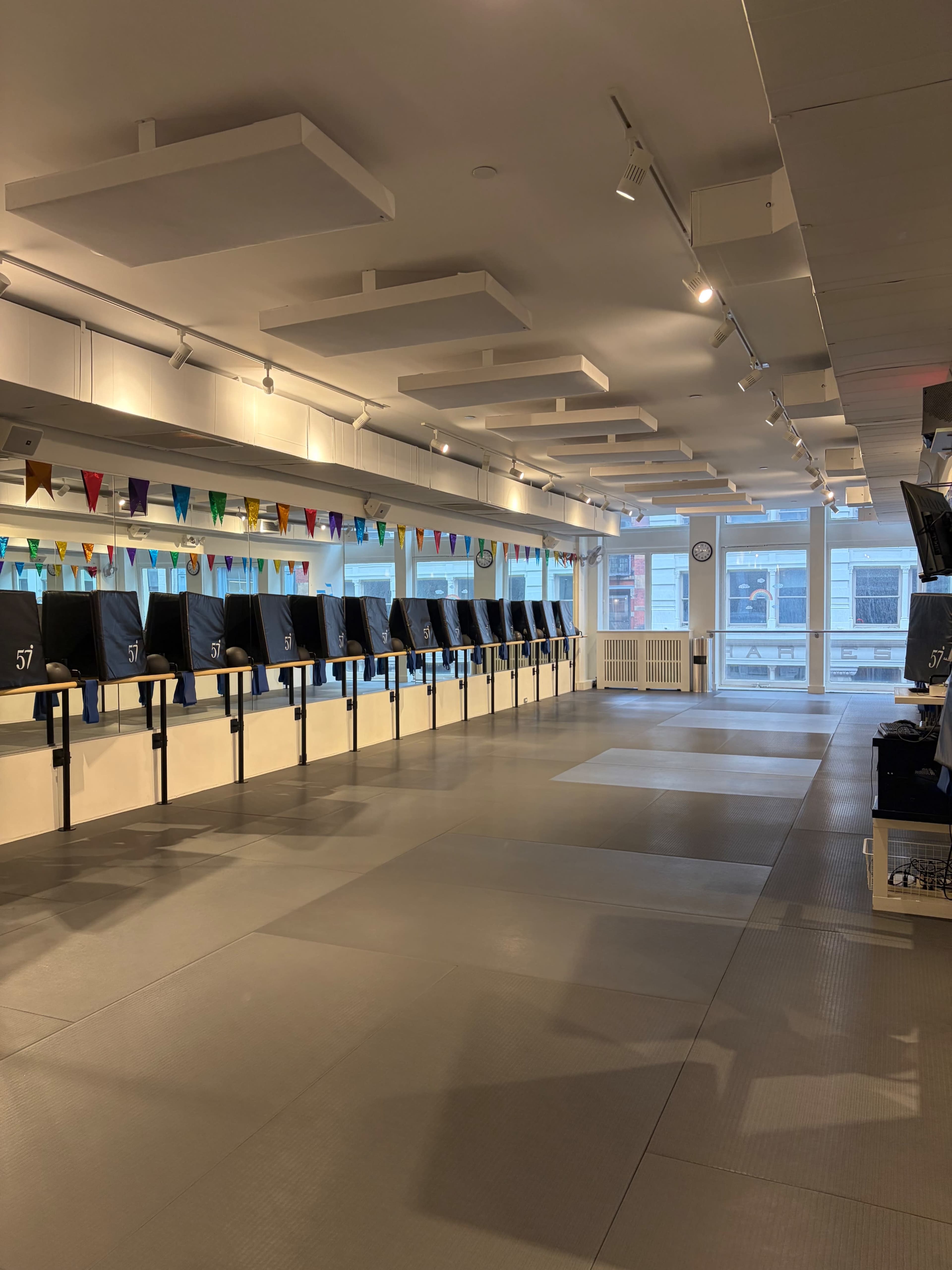 The image shows a spacious, vacant fitness studio with mirrored walls, exercise equipment lined up, and colorful bunting hanging from the ceiling.
