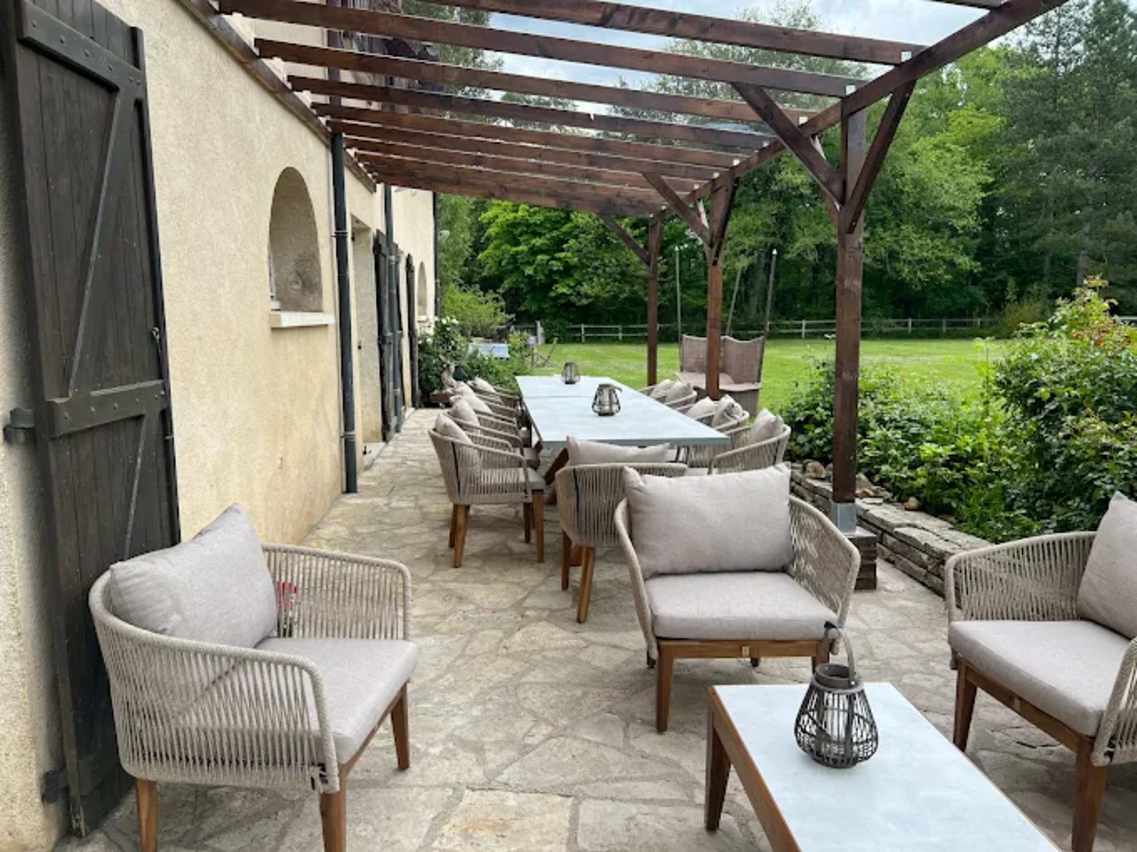 A shaded patio features wooden frames and a long table surrounded by several upholstered chairs, with greenery visible in the background.
