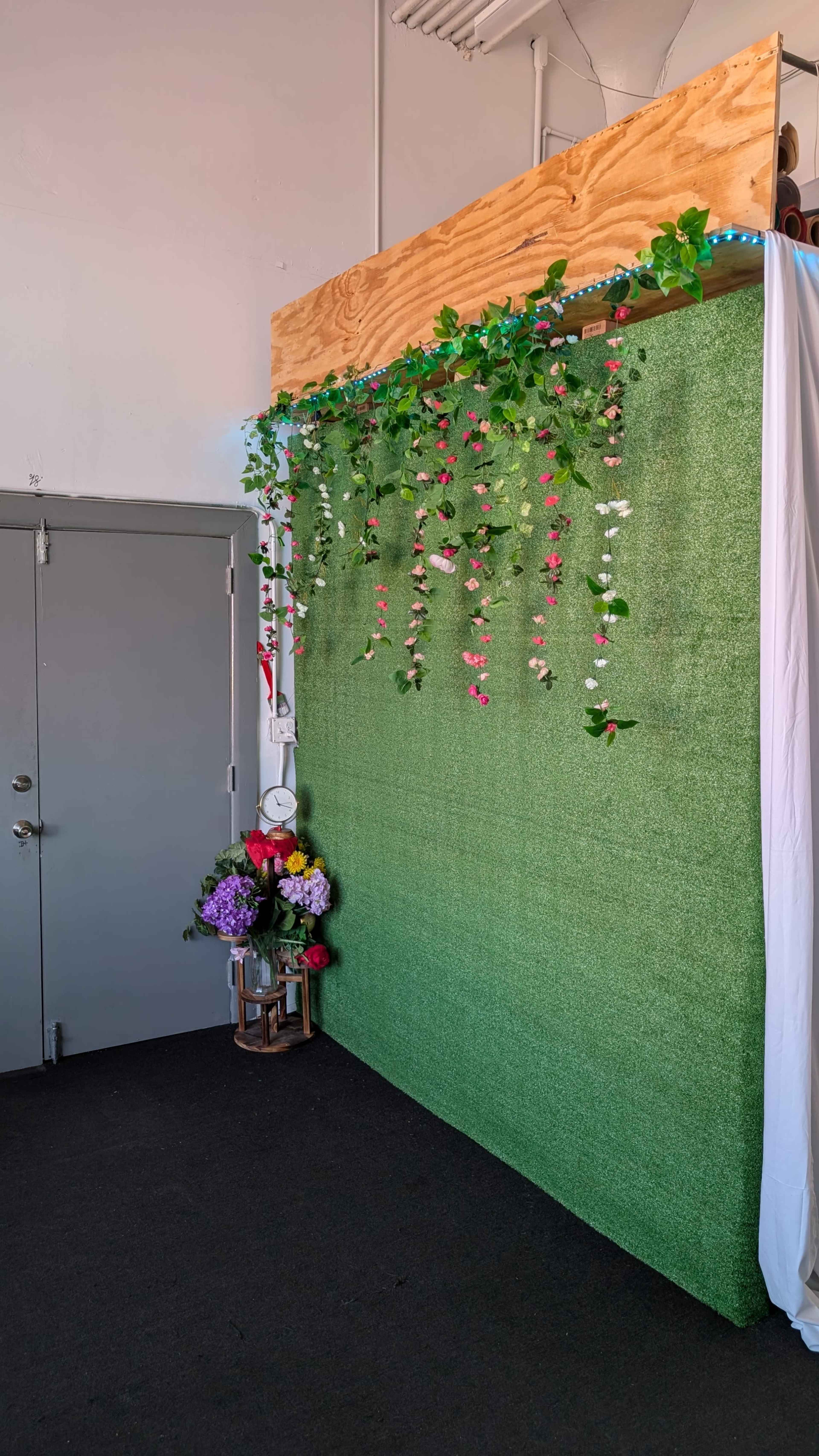 The image shows a green backdrop adorned with hanging artificial flowers and foliage, alongside a corner featuring a bouquet of colorful flowers in a lantern.