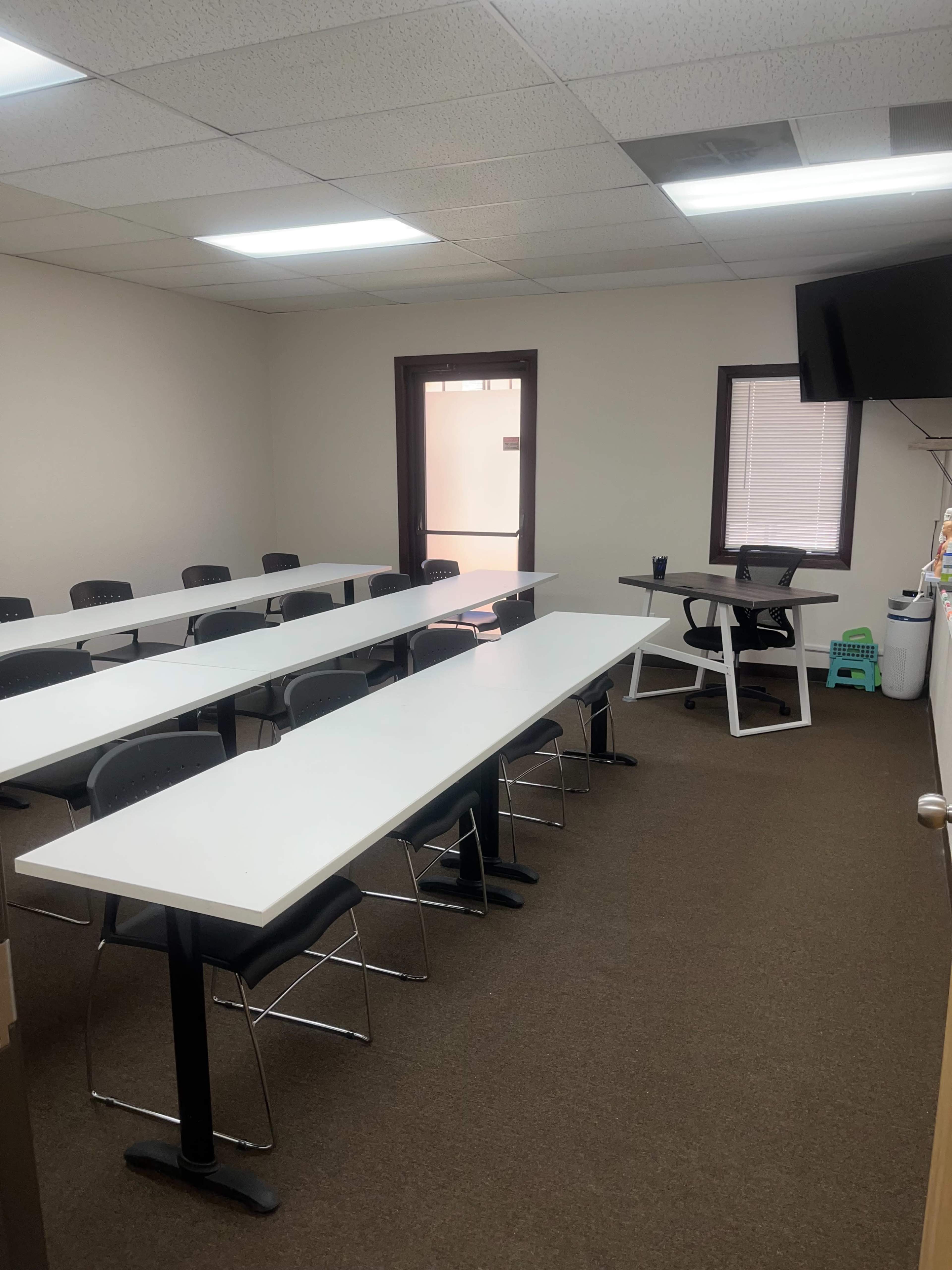 The image shows a meeting room with several white tables arranged in a U-shape, equipped with black chairs and a television screen on one wall.
