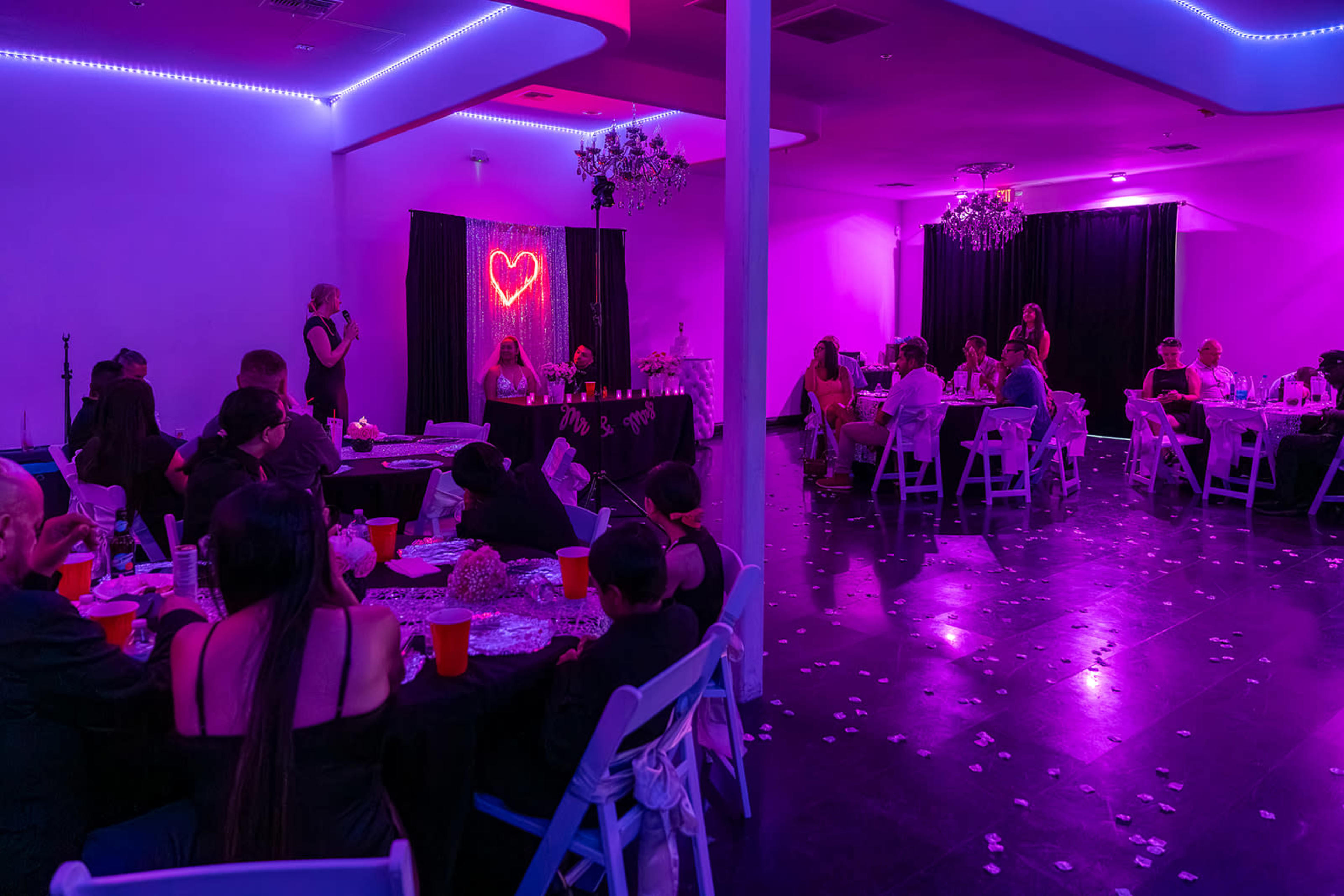 A decorated event space features guests seated at tables under vibrant purple lighting, with a performer speaking on stage next to a heart-shaped neon sign.