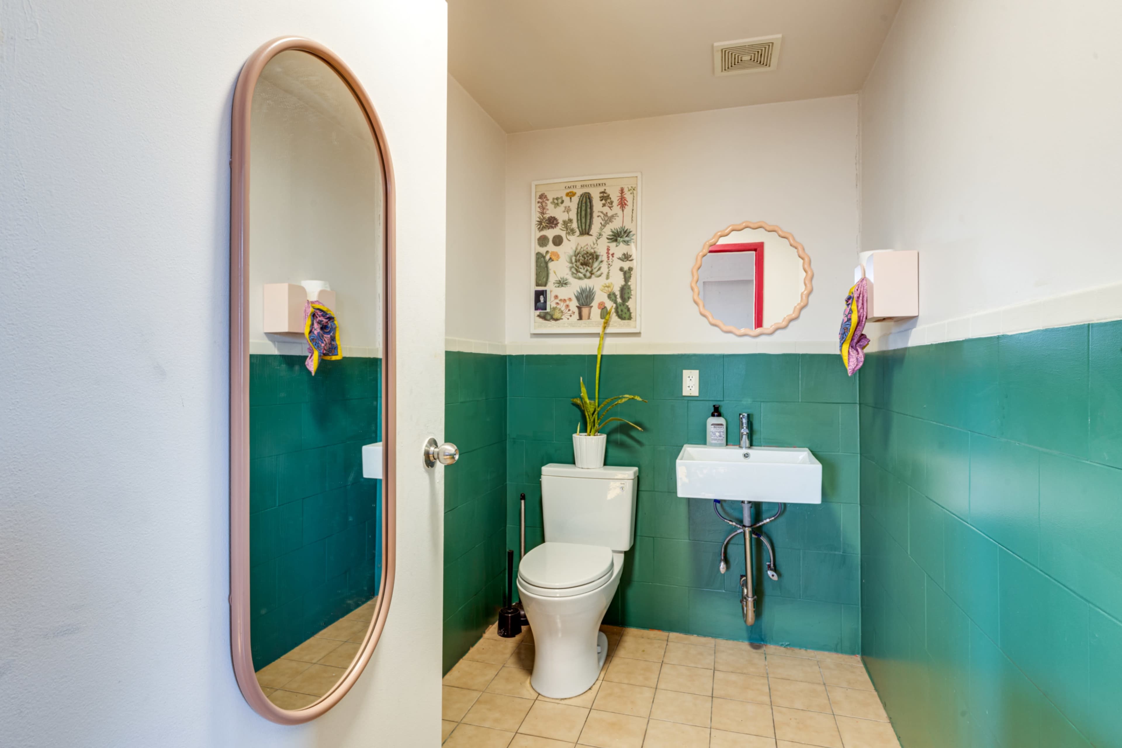 A small bathroom features a green tiled wall, a white toilet, a pedestal sink, and a round mirror with a pink frame.