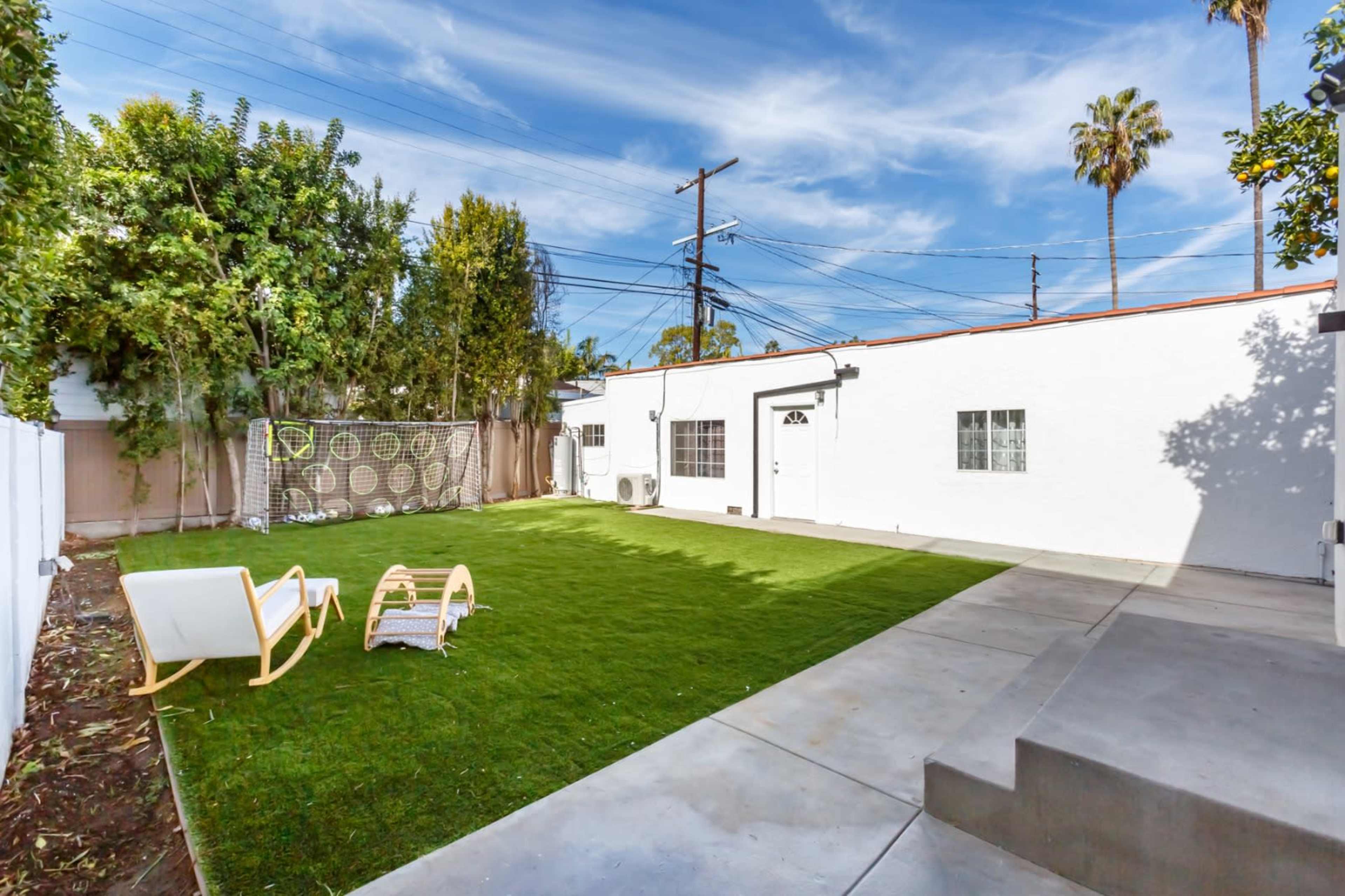 A spacious backyard features a green lawn, a seating area with two chairs, and a concrete patio adjacent to a white house.