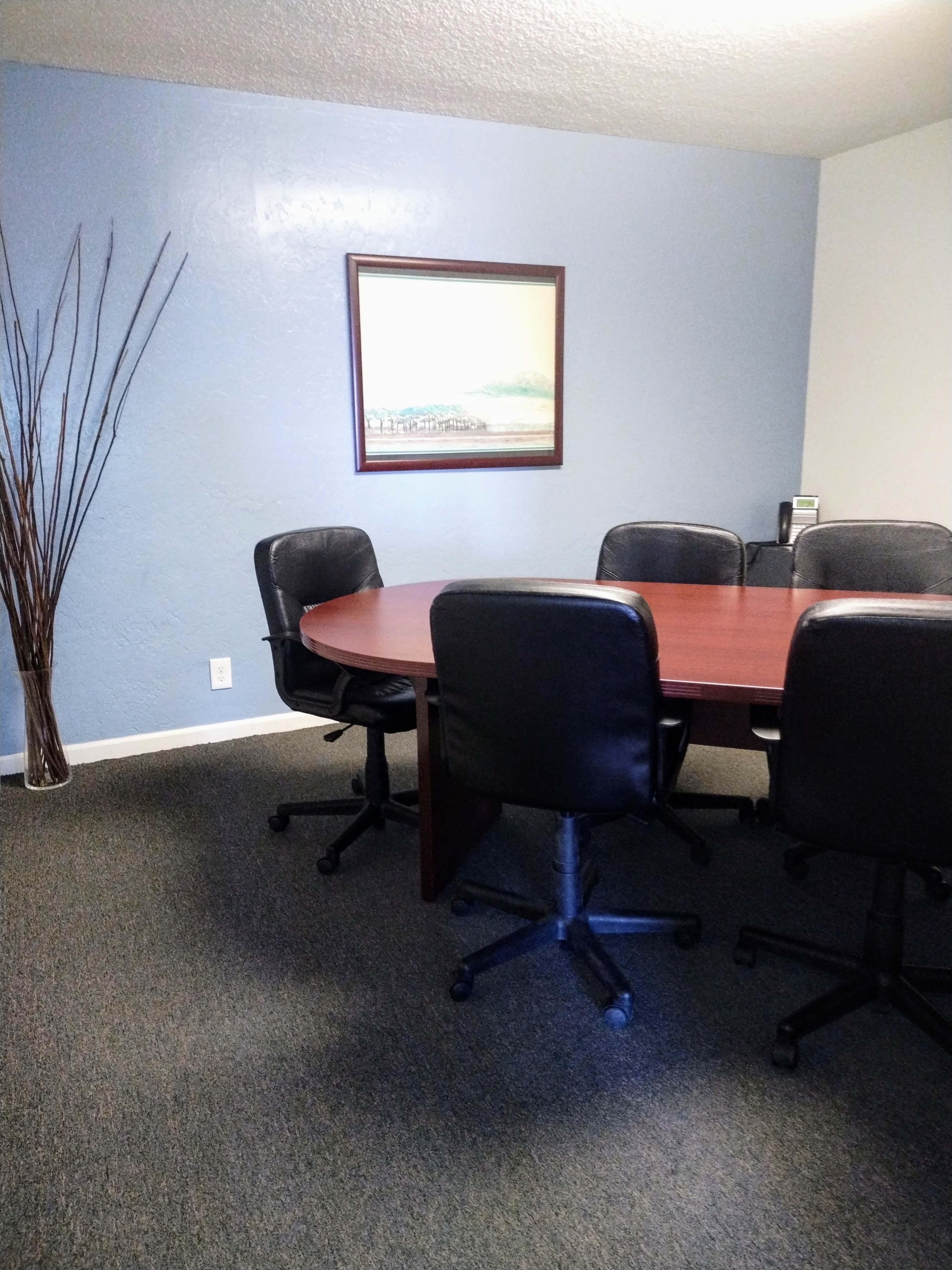 A conference room is shown with a large circular table surrounded by black chairs and a wall with a painting.