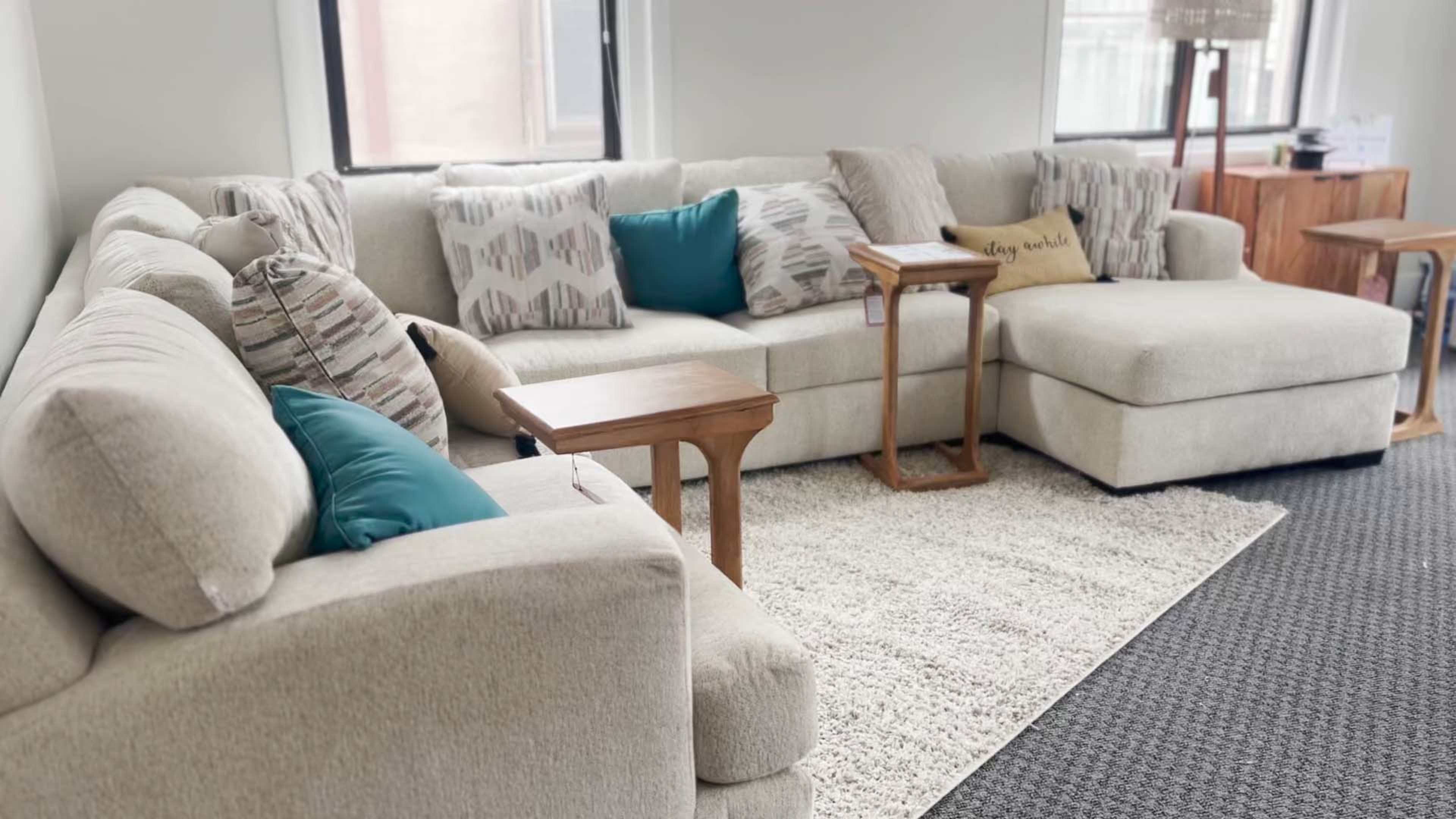 A light-colored sectional sofa adorned with multiple decorative pillows, positioned on a textured rug in a bright room with windows.
