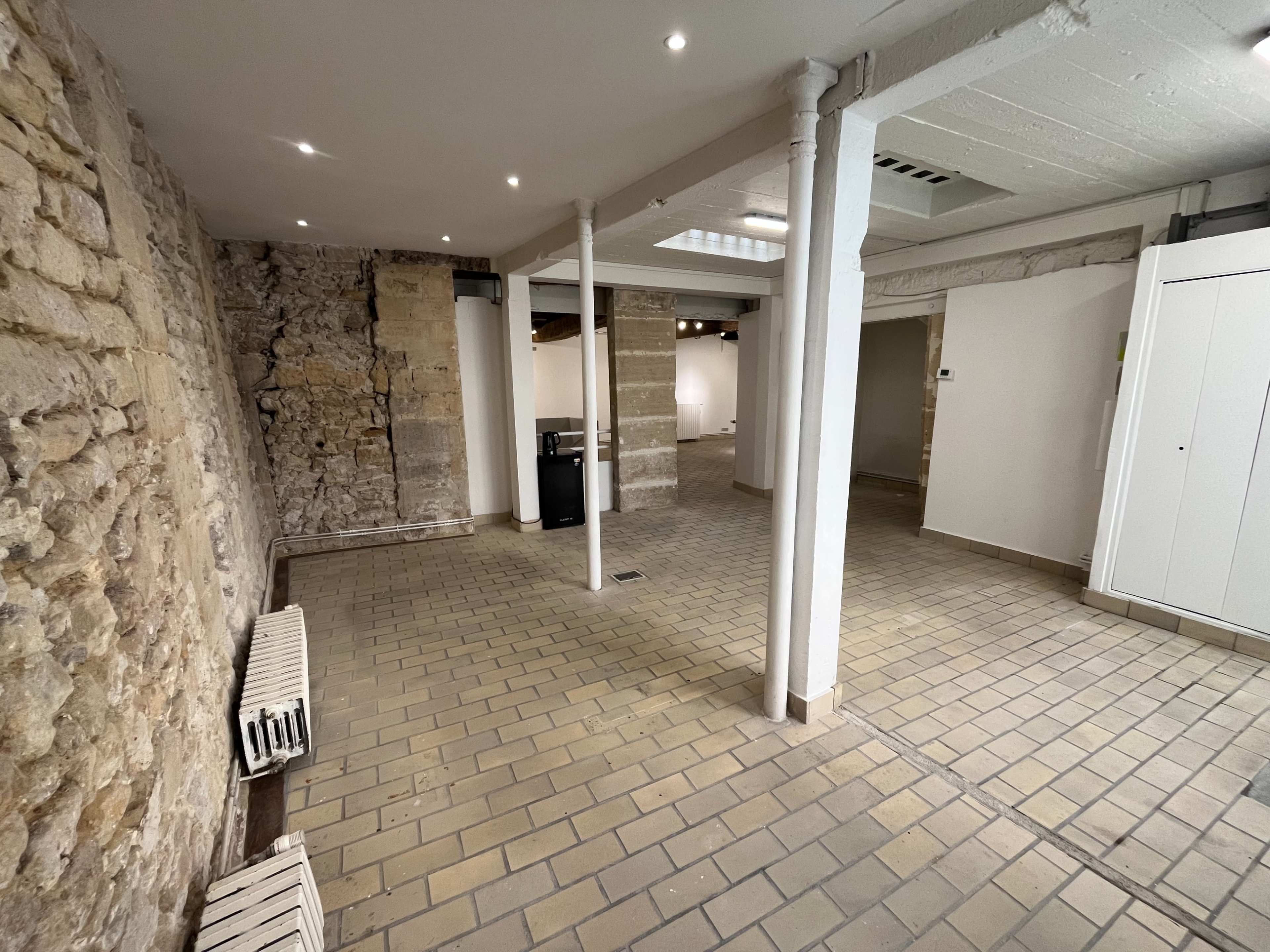 The image depicts a spacious room with a stone wall on one side, tiled flooring, and several structural columns supporting the ceiling.