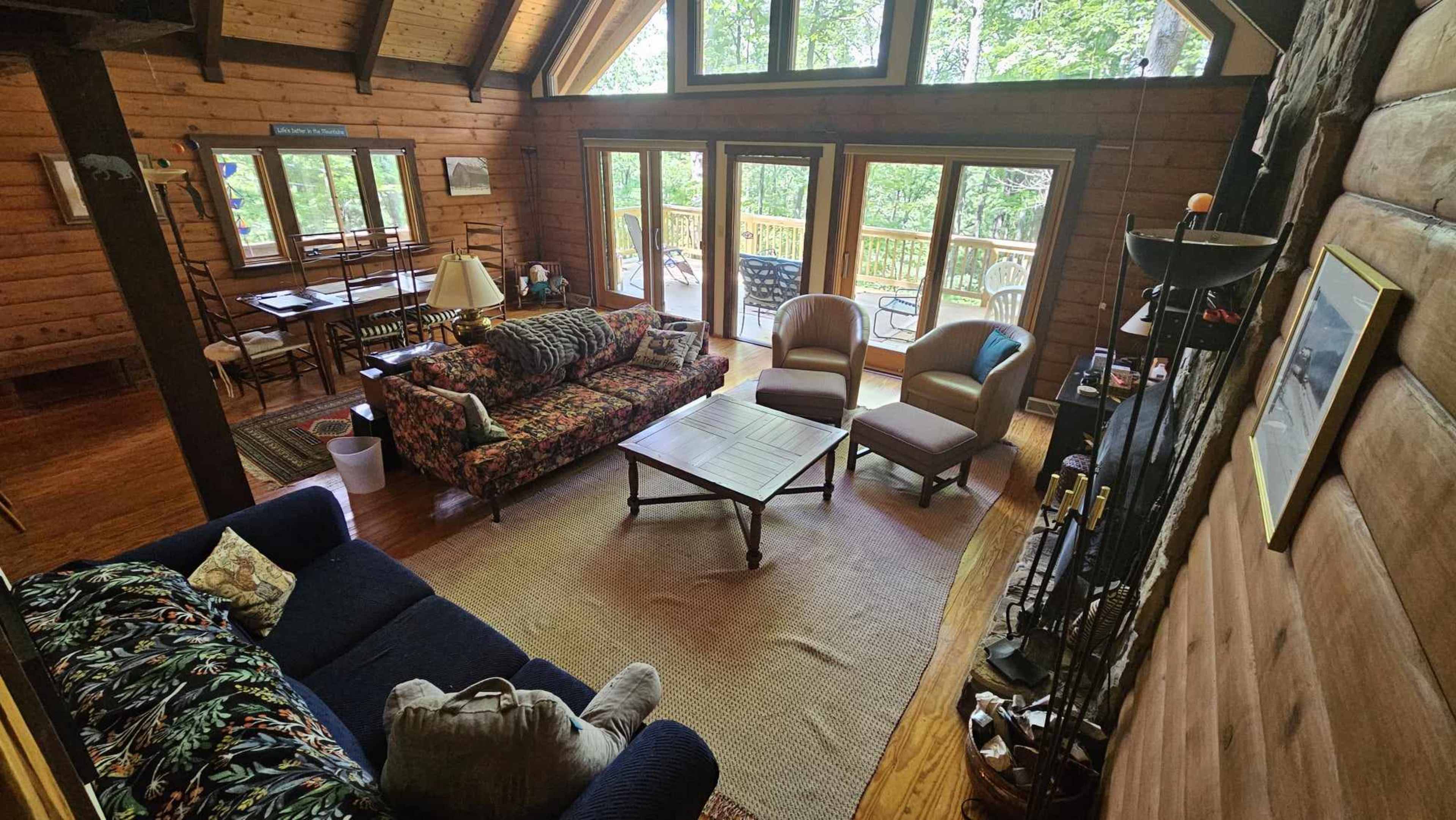 The image shows a cozy cabin interior with wooden walls, a seating area featuring floral-patterned sofas, and large windows overlooking a forested deck.