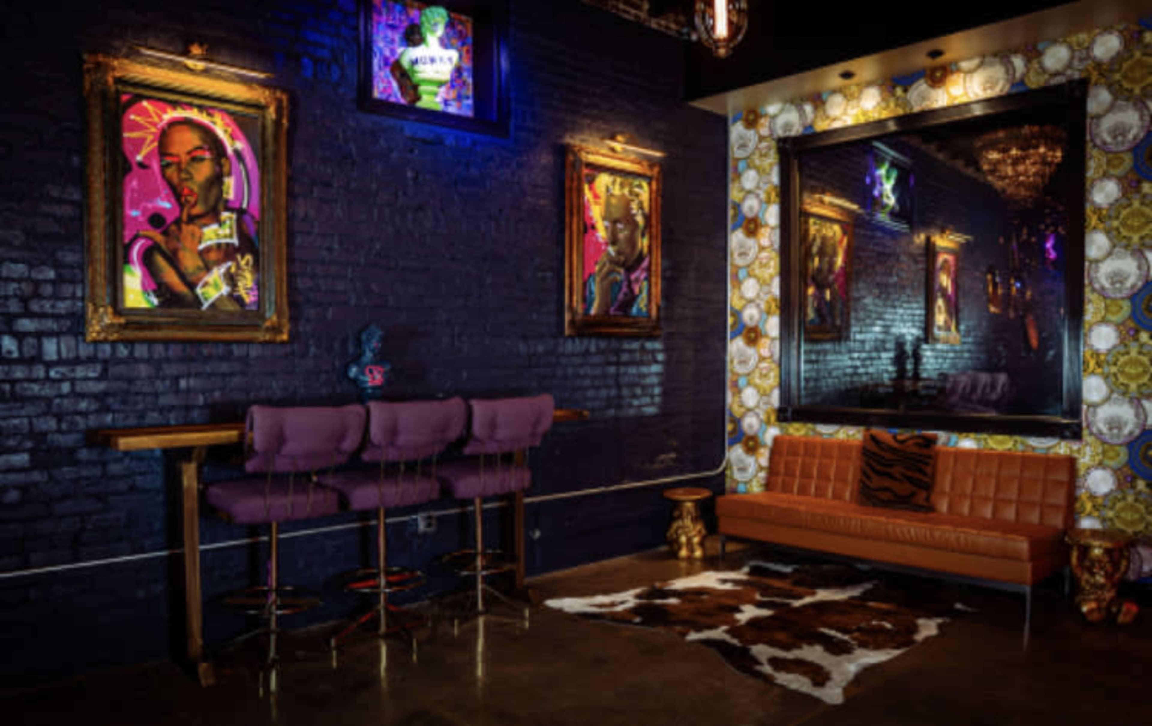 A stylish interior featuring purple bar stools, an orange couch with a tiger-patterned pillow, colorful framed artwork on dark walls, and a large mirror surrounded by ornate wallpaper.