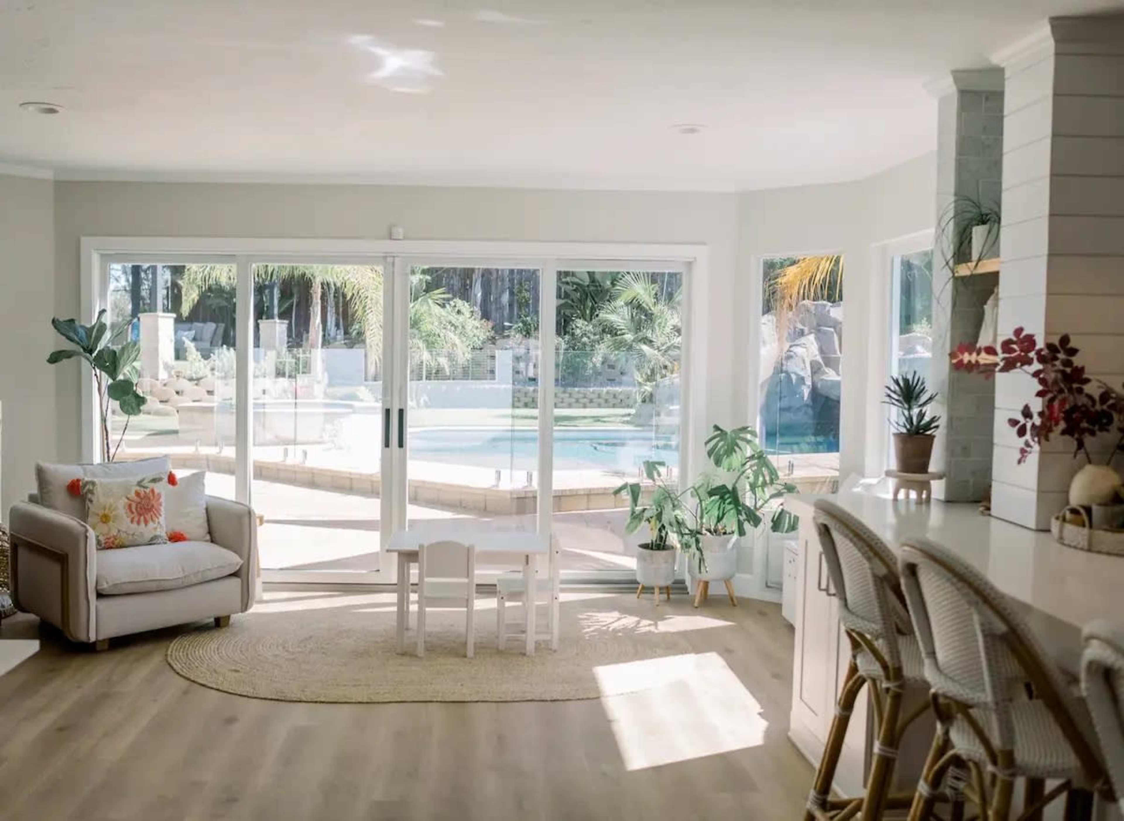 A bright living room features large glass sliding doors overlooking a pool area, with a small table and chairs in the center and plants scattered around.