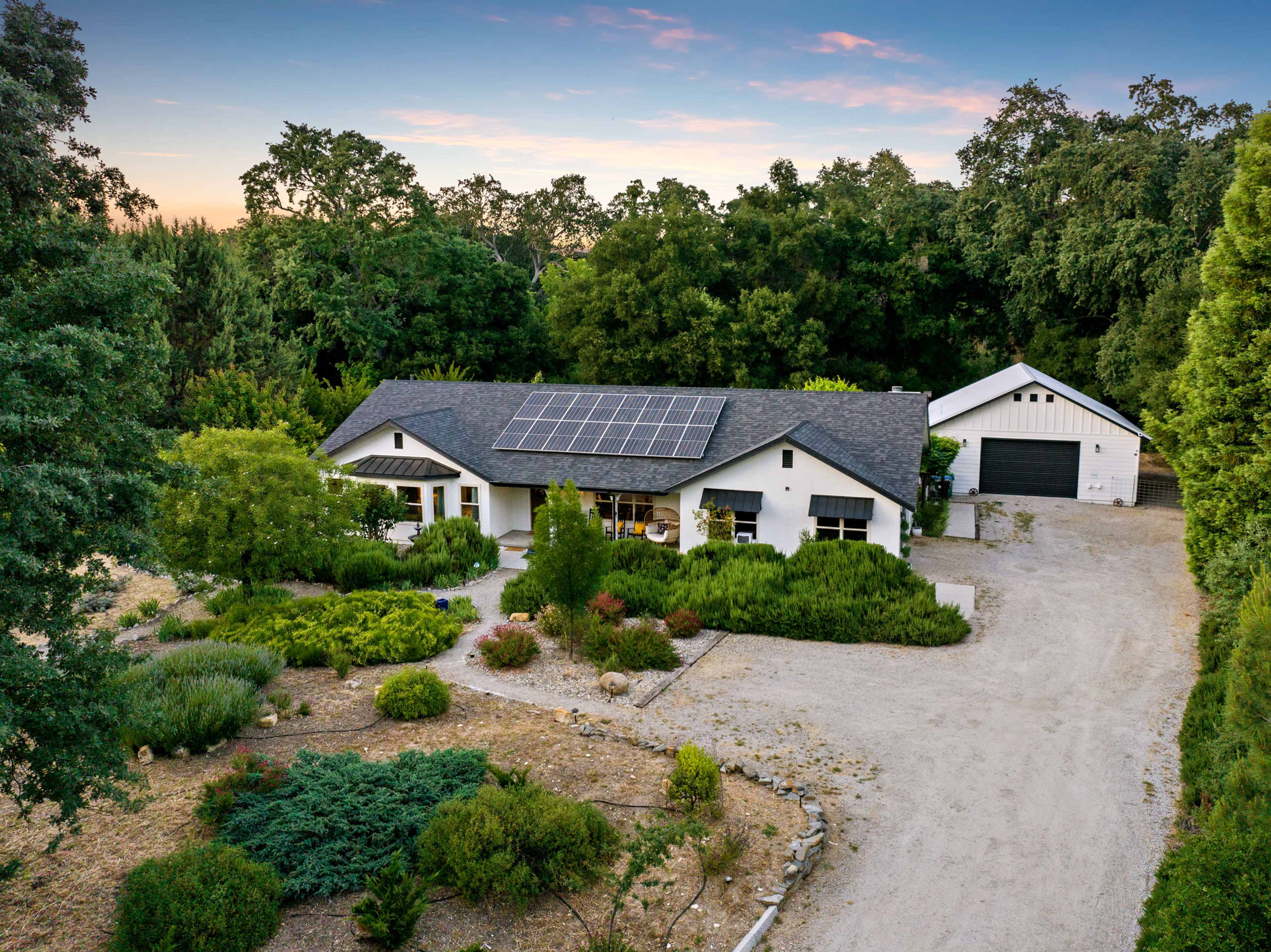 A modern house with solar panels on the roof is surrounded by landscaped greenery and a gravel driveway, leading to a separate garage.