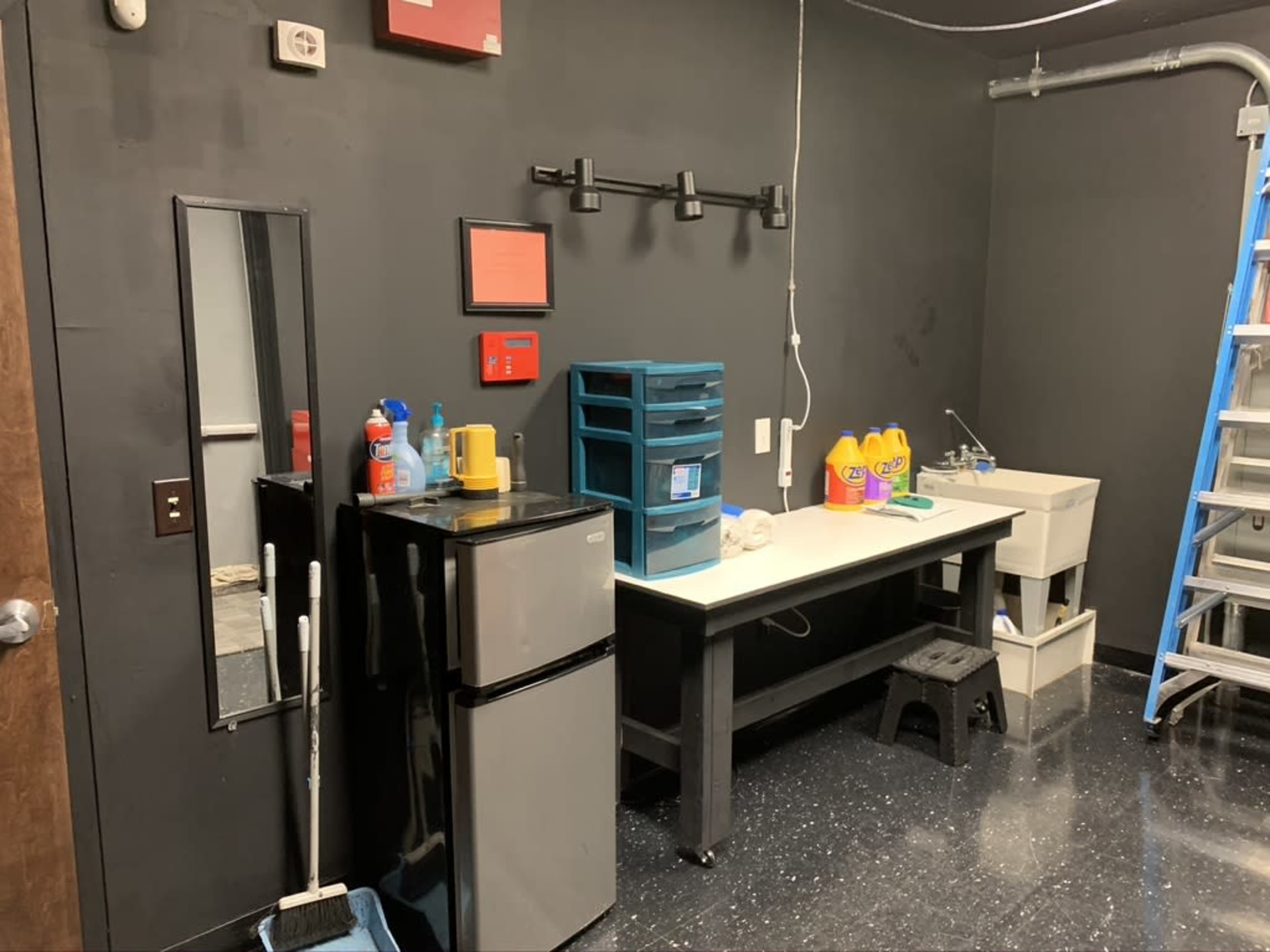 A small utility room with a black wall, a refrigerator, a table with cleaning supplies, and a sink, along with a ladder in the corner.