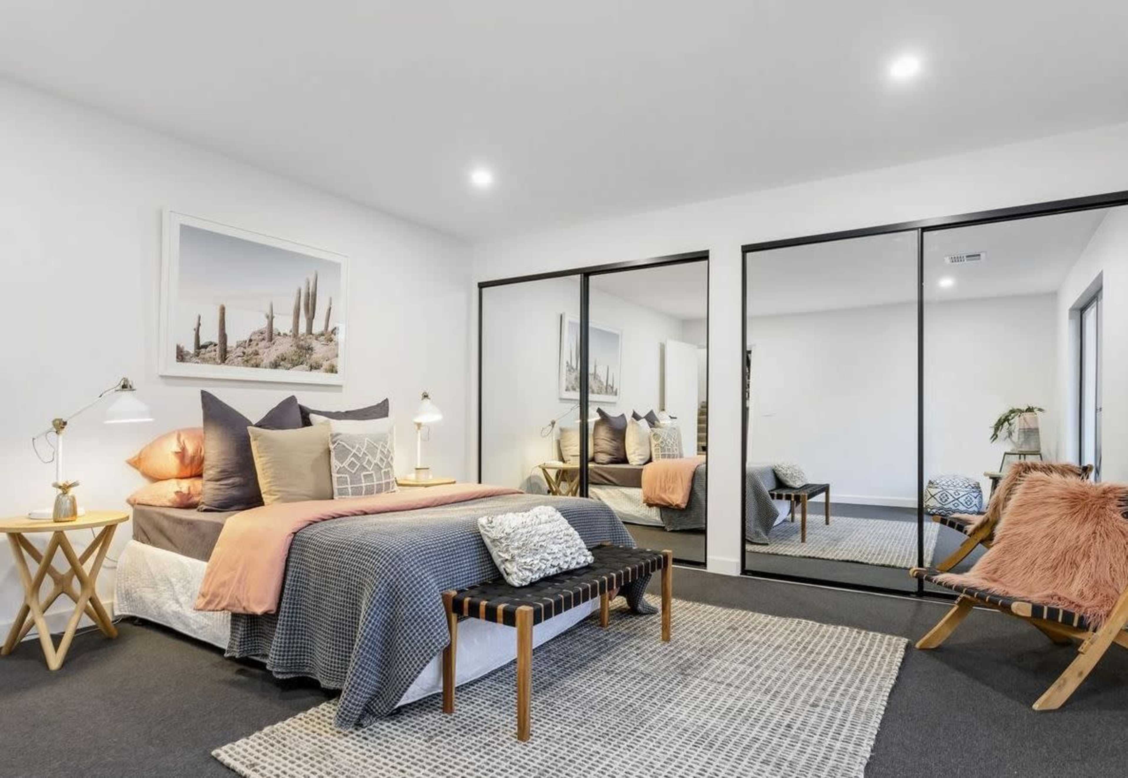 A modern bedroom with a bed covered in various textured pillows, a mirror wardrobe, a small bench at the foot of the bed, and artwork on the wall.