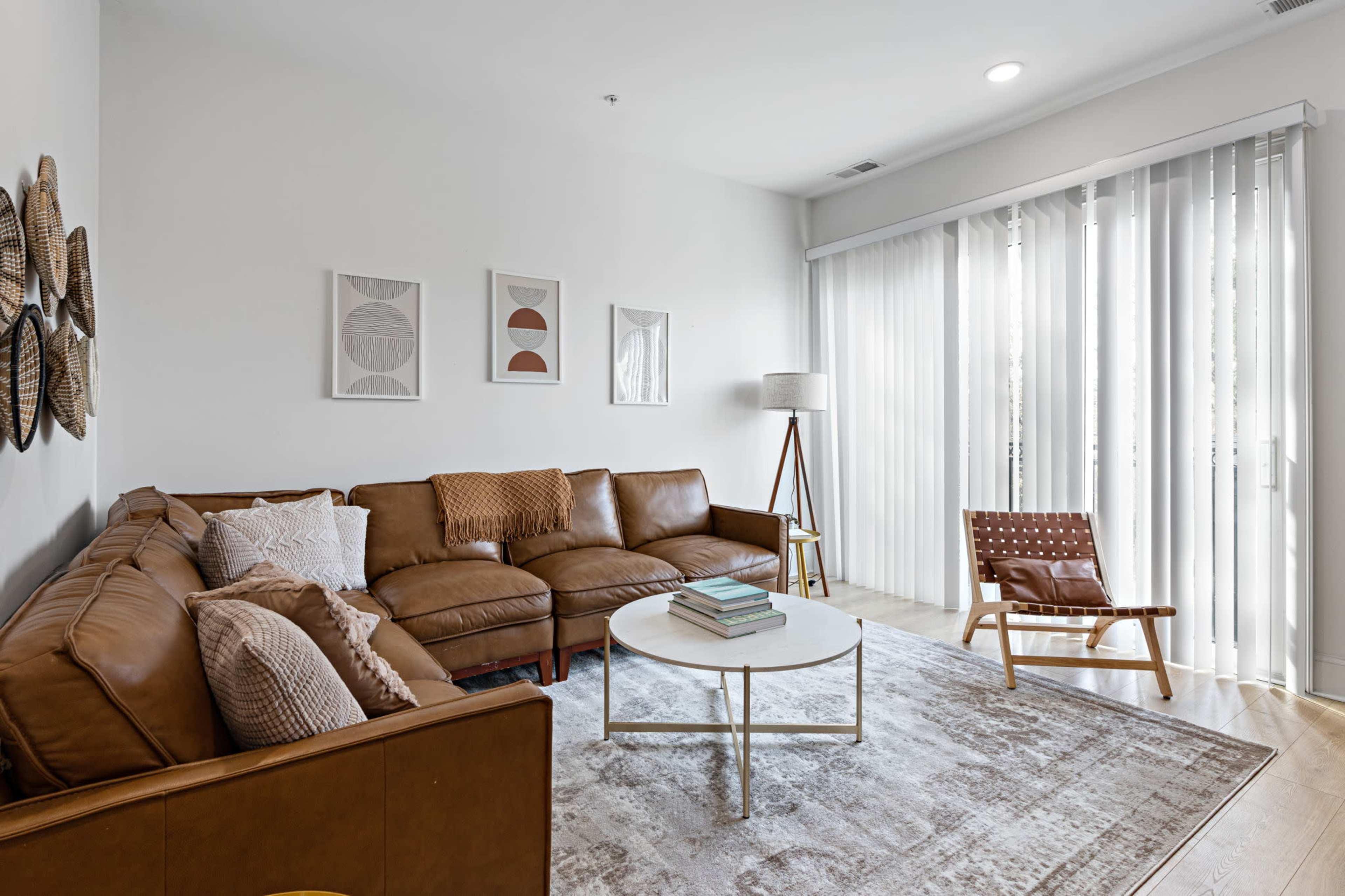 The image shows a modern living room featuring a brown leather sectional sofa, a round white coffee table, a standing lamp, and large windows covered with sheer curtains.