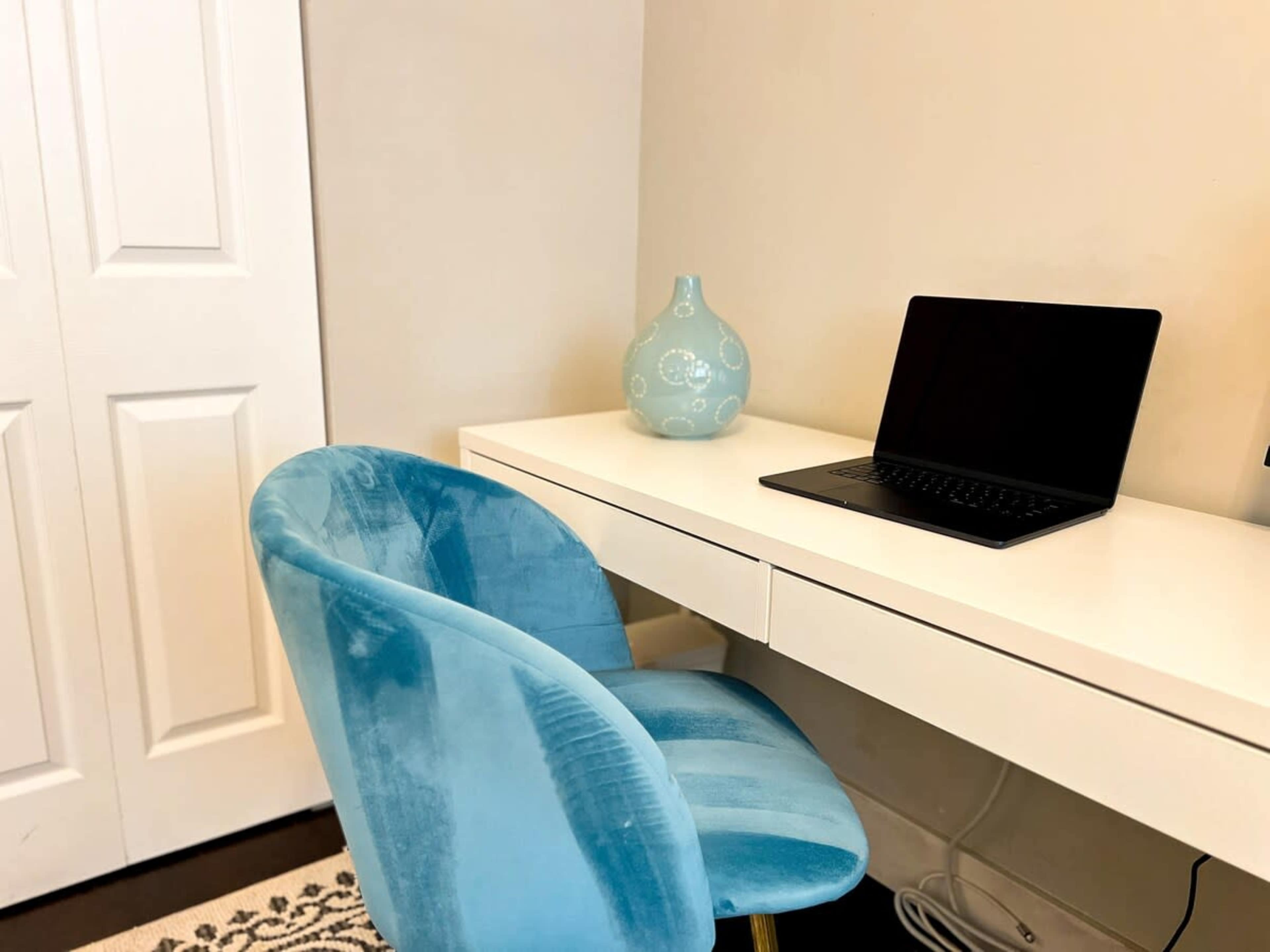 A blue velvet chair is positioned in front of a white desk with a closed laptop and a decorative blue vase beside it, against a light-colored wall and a closet door.
