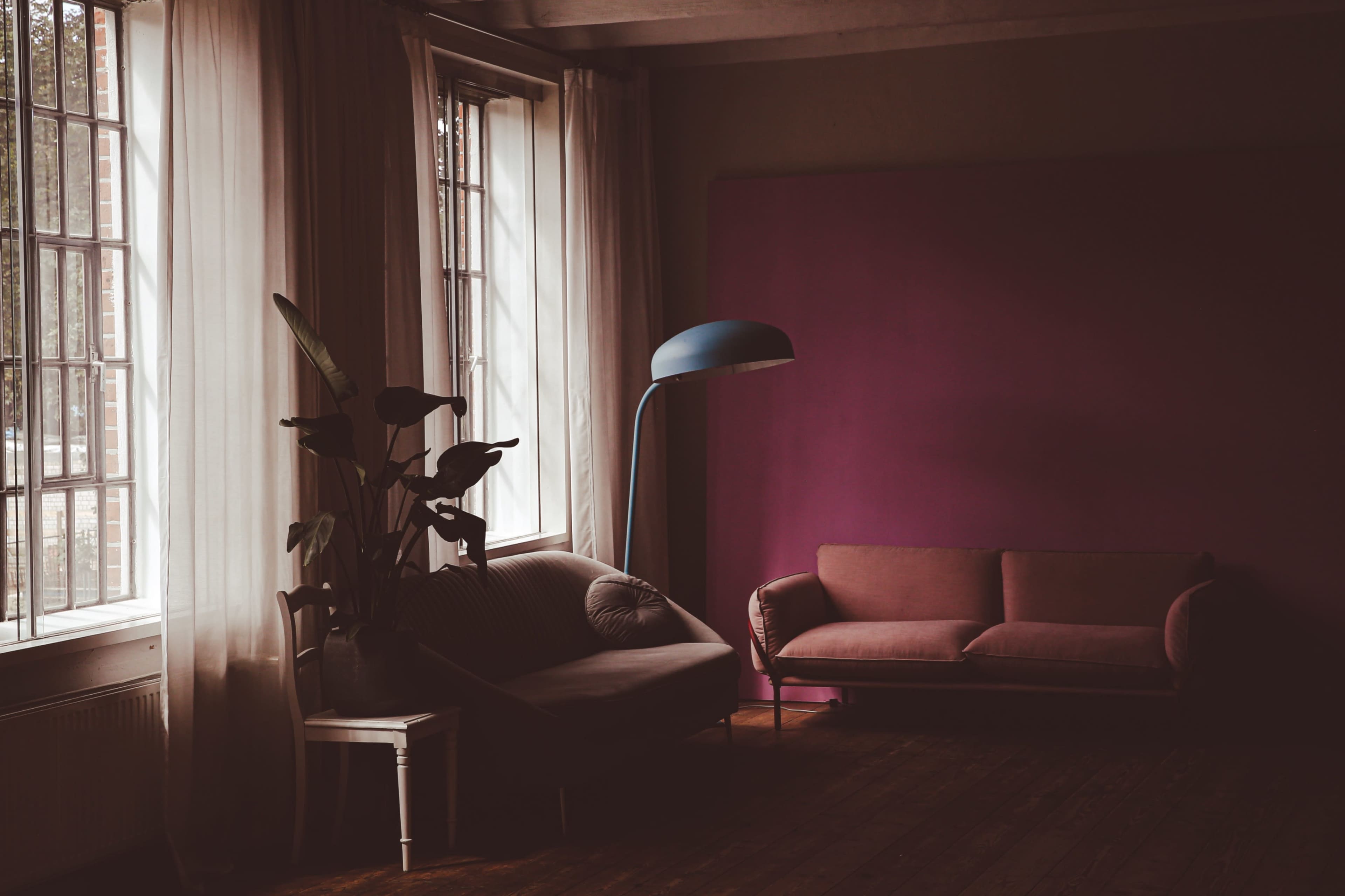 A room features two sofas, a floor lamp, and large windows draped with sheer curtains, creating a serene atmosphere with a purple wall backdrop.