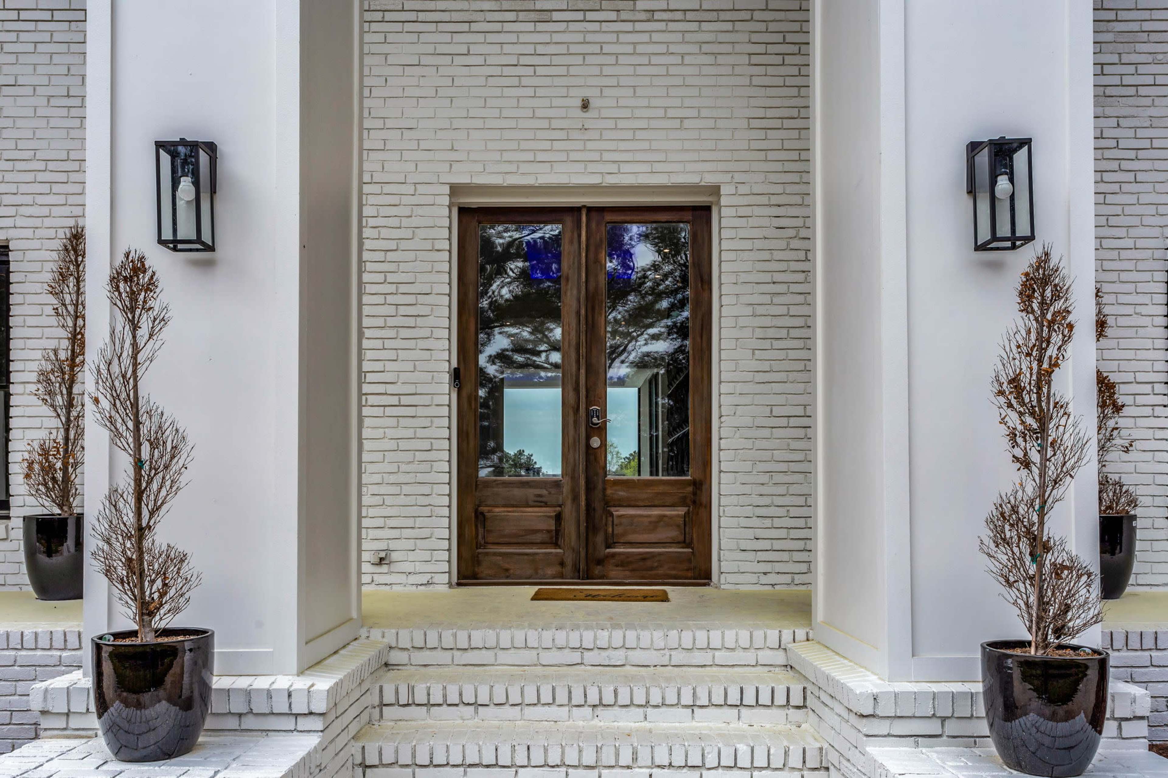 A symmetrical entrance features double wooden doors flanked by tall potted shrubs and white brick walls.