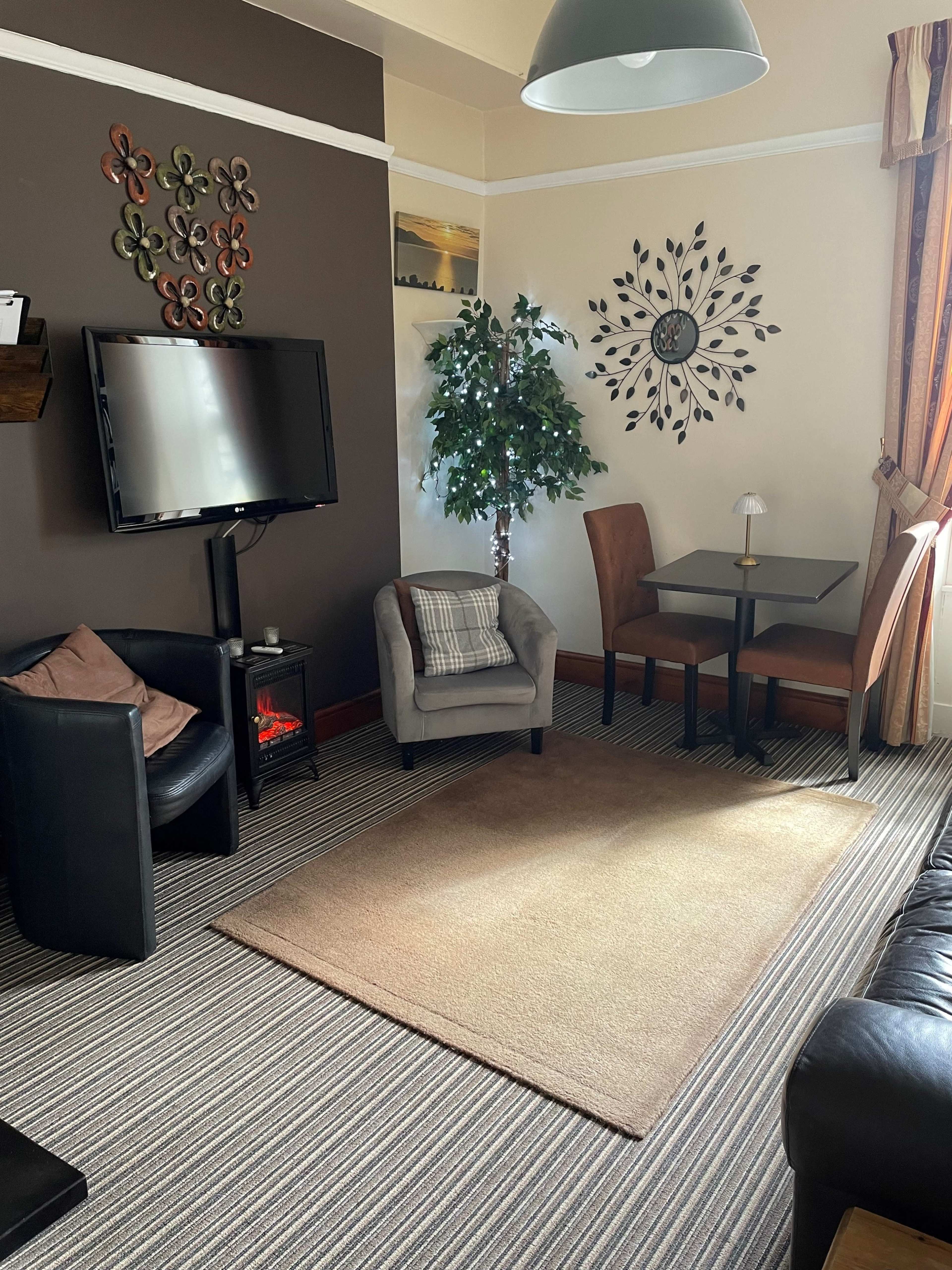 The living room features a television, a small dining table with chairs, a faux tree, and a patterned rug on striped flooring.