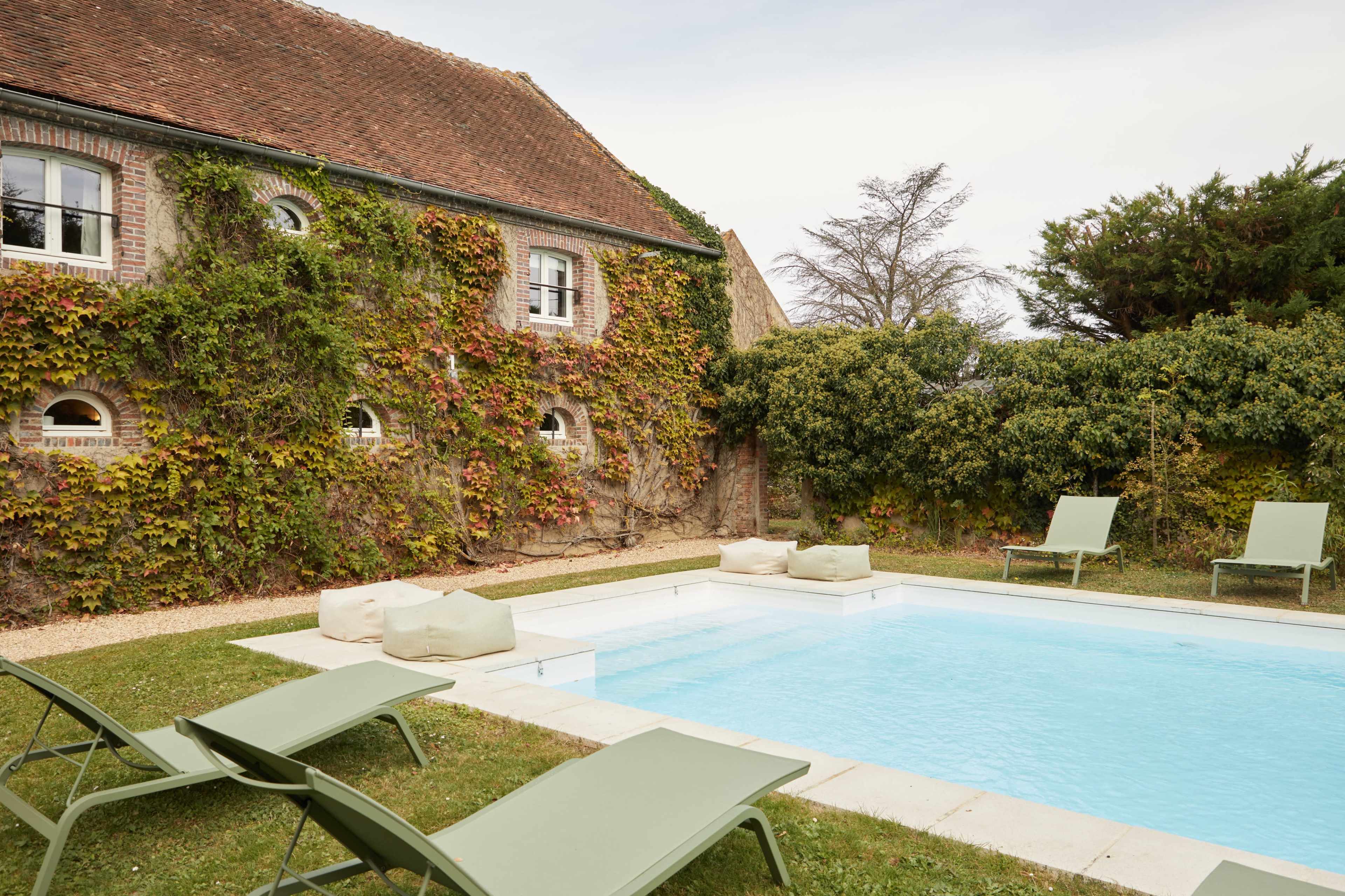 A rectangular swimming pool is surrounded by lounge chairs and lush greenery next to a brick building covered in ivy.