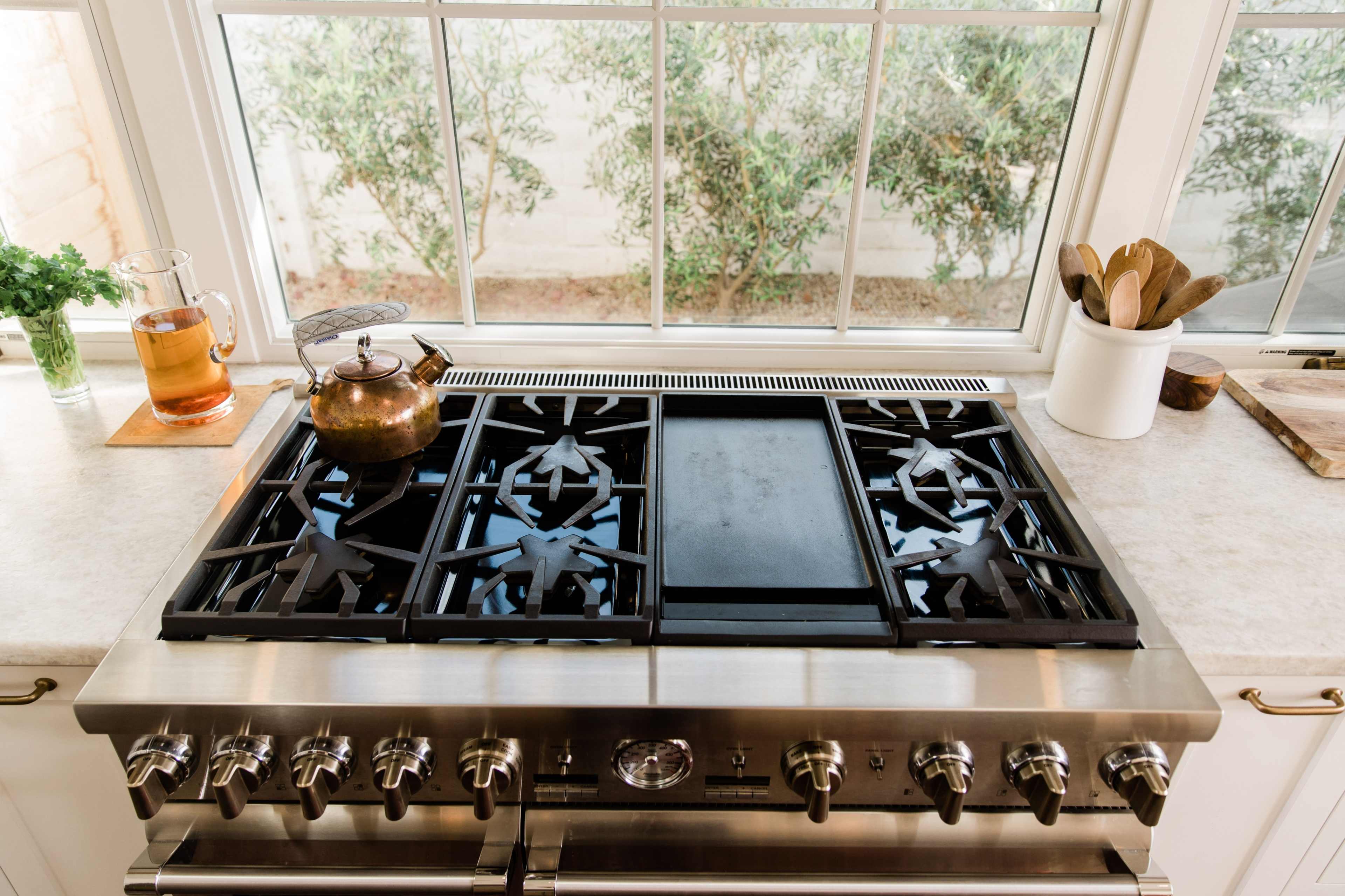 The image shows a modern kitchen stove with a visible gas burner and griddle, set on a countertop near a window with green plants outside.
