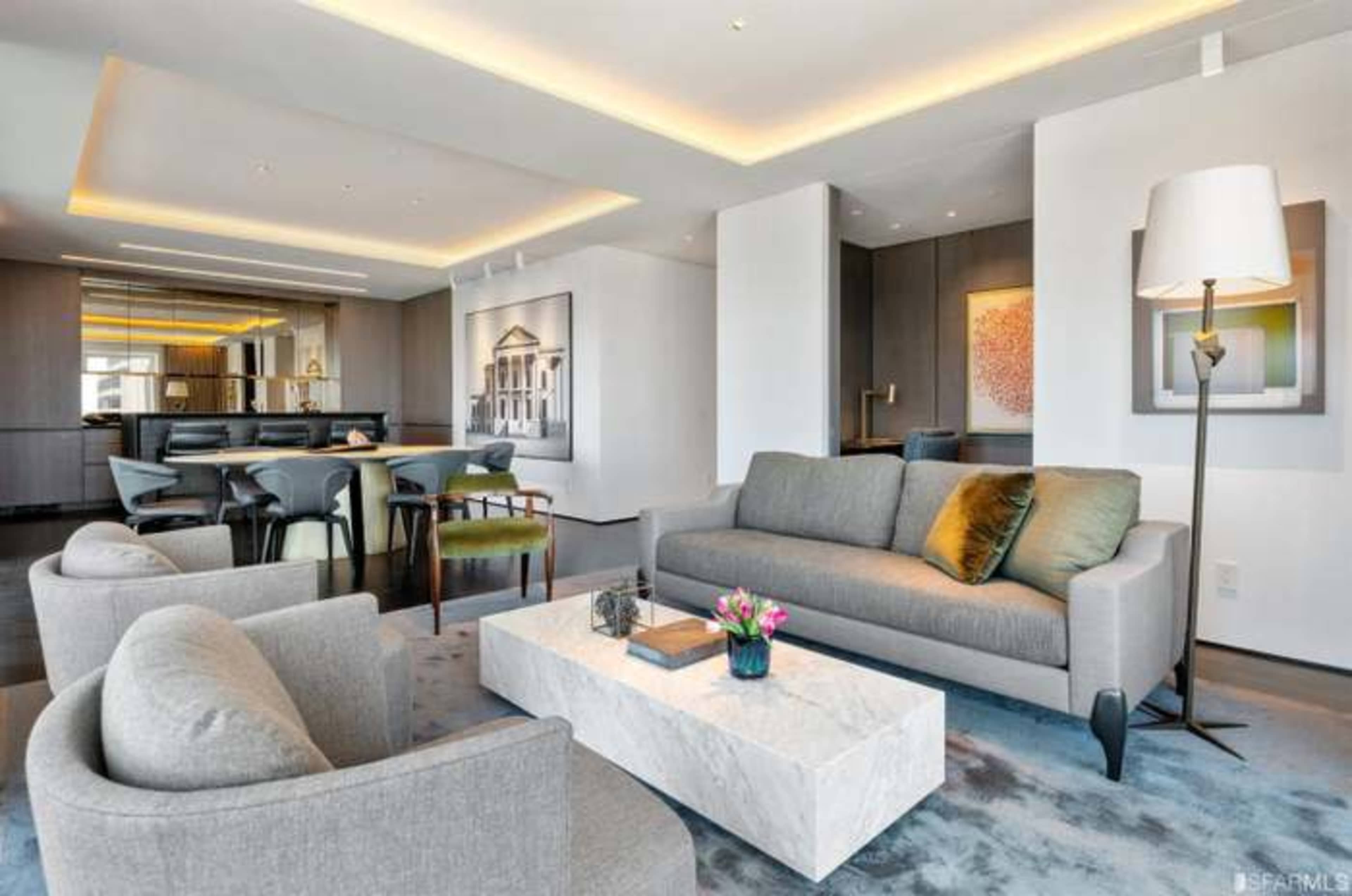 A modern living room with a gray sofa, a marble coffee table, and seating around a dining area in the background.